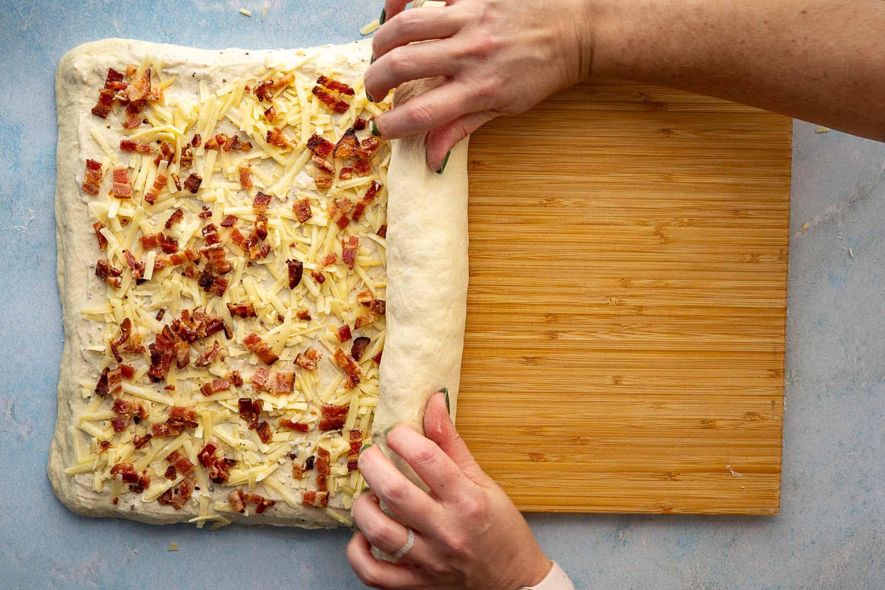 Hands rolling up dough topped with shredded cheese and bacon bits on a wooden board, preparing to make a filled bread or savory roll.
