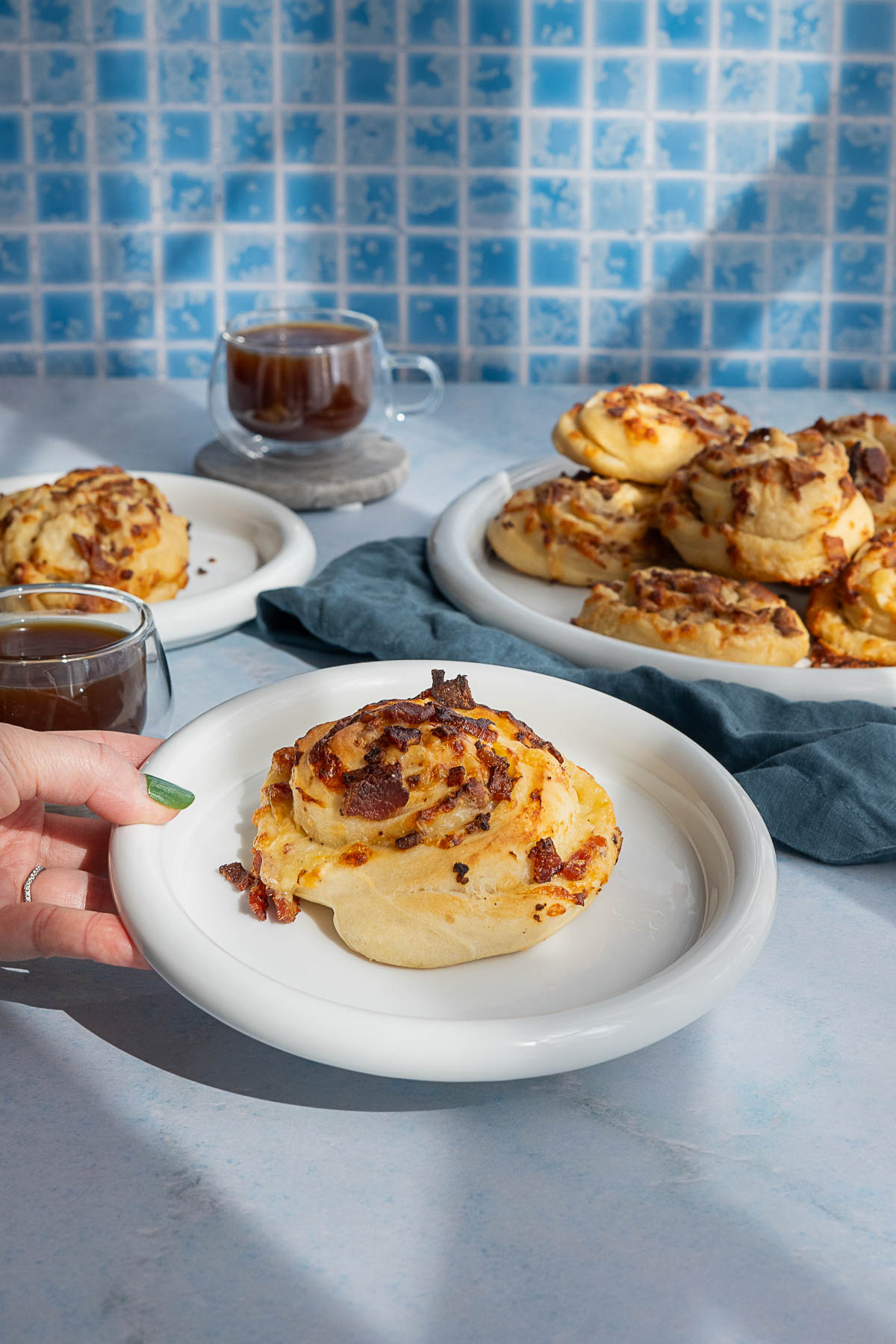 A hand holds a plate with a swirl-shaped pastry topped with bacon, with more pastries and cups of coffee in the background on a blue counter and tile backdrop.