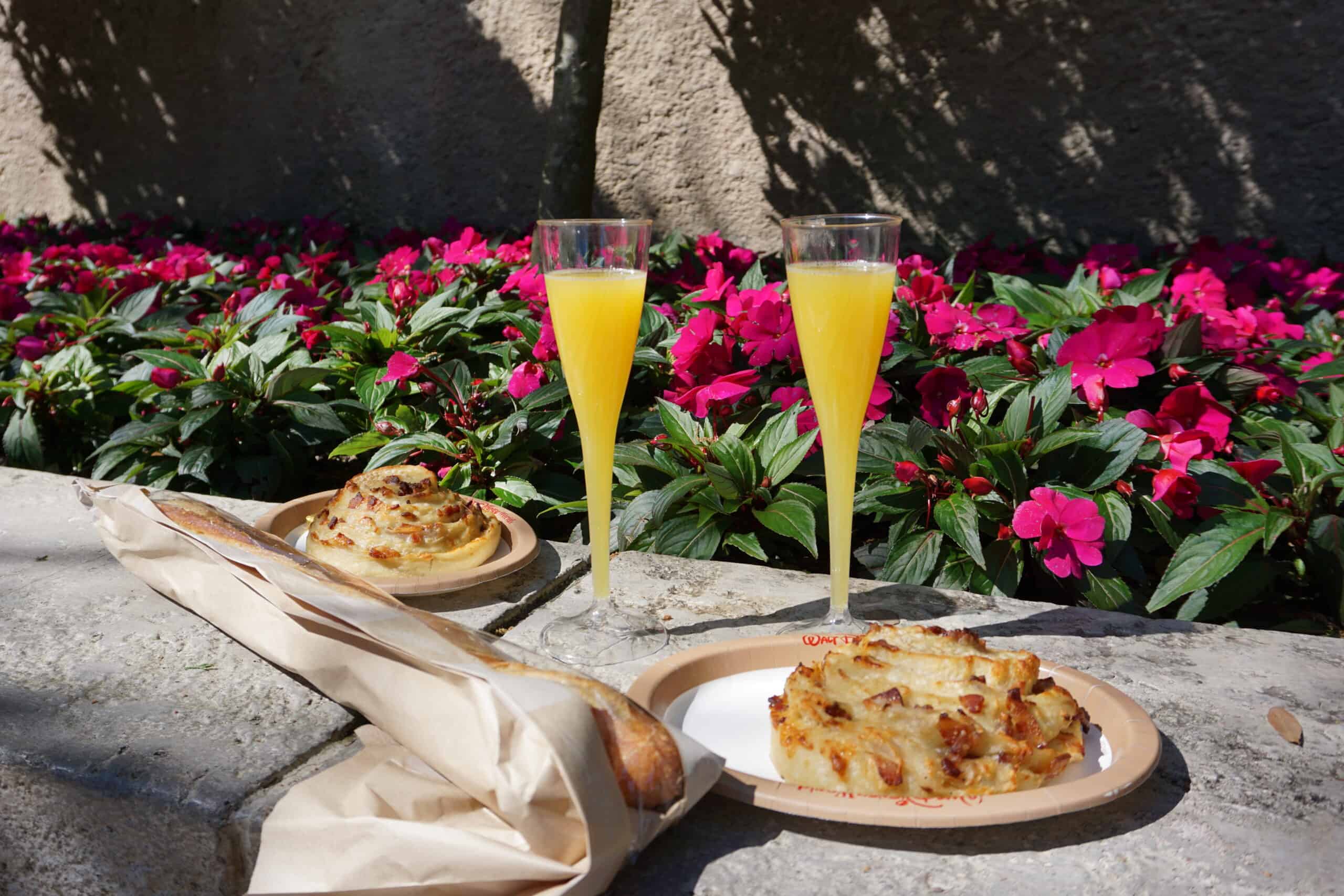 Two glasses of orange juice, two bacon and cheese rolls on plates, and a baguette laid out on a stone surface, with pink flowers and green leaves in the background.
