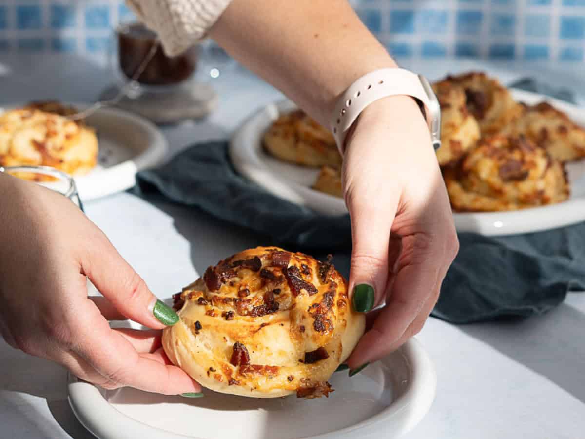 A person with green nail polish places a bacon and cheese roll onto a white plate. More bacon and cheese rolls are visible in the background on a tray as sunlight casts shadows across the table.
