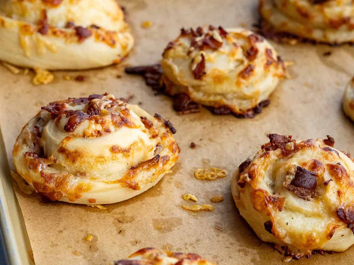 Golden brown bacon and cheese rolls rest on a parchment-lined baking sheet, their swirled dough revealing melted cheese and crispy bacon bits for a savory treat.