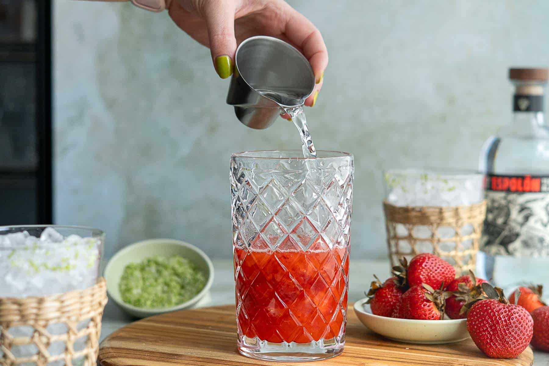 A hand pours clear liquid from a metal jigger into a glass with red juice on a wooden board; nearby are fresh strawberries, a bowl of green zest, and ice-filled glasses—perfect for crafting a strawberry margarita on the rocks.