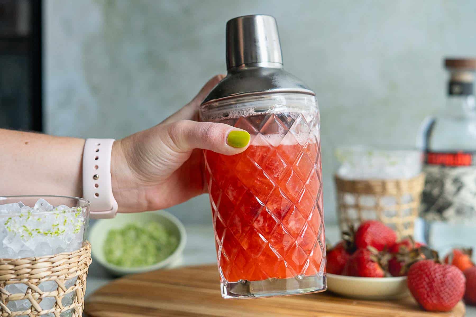 A hand with green painted nails holds a cocktail shaker filled with a bright red drink. Nearby are fresh strawberries, a glass of ice for your strawberry margarita on the rocks, a bowl of green ingredient, and a bottle of liquor on a wooden surface.