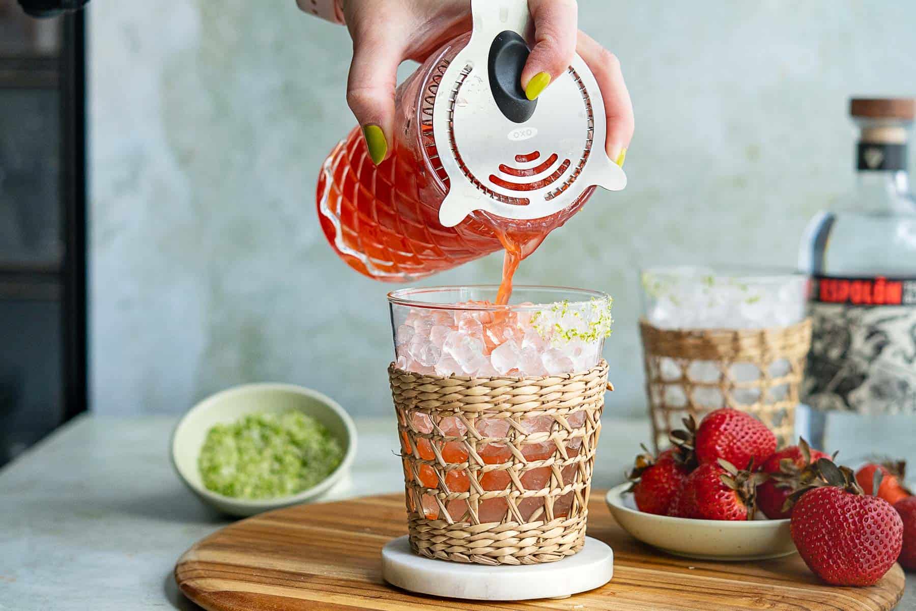 A hand pours a vibrant strawberry margarita on the rocks through a strainer into an ice-filled glass with a woven sleeve. Fresh strawberries and a bowl of green salt or seasoning rest nearby on a wooden board.
