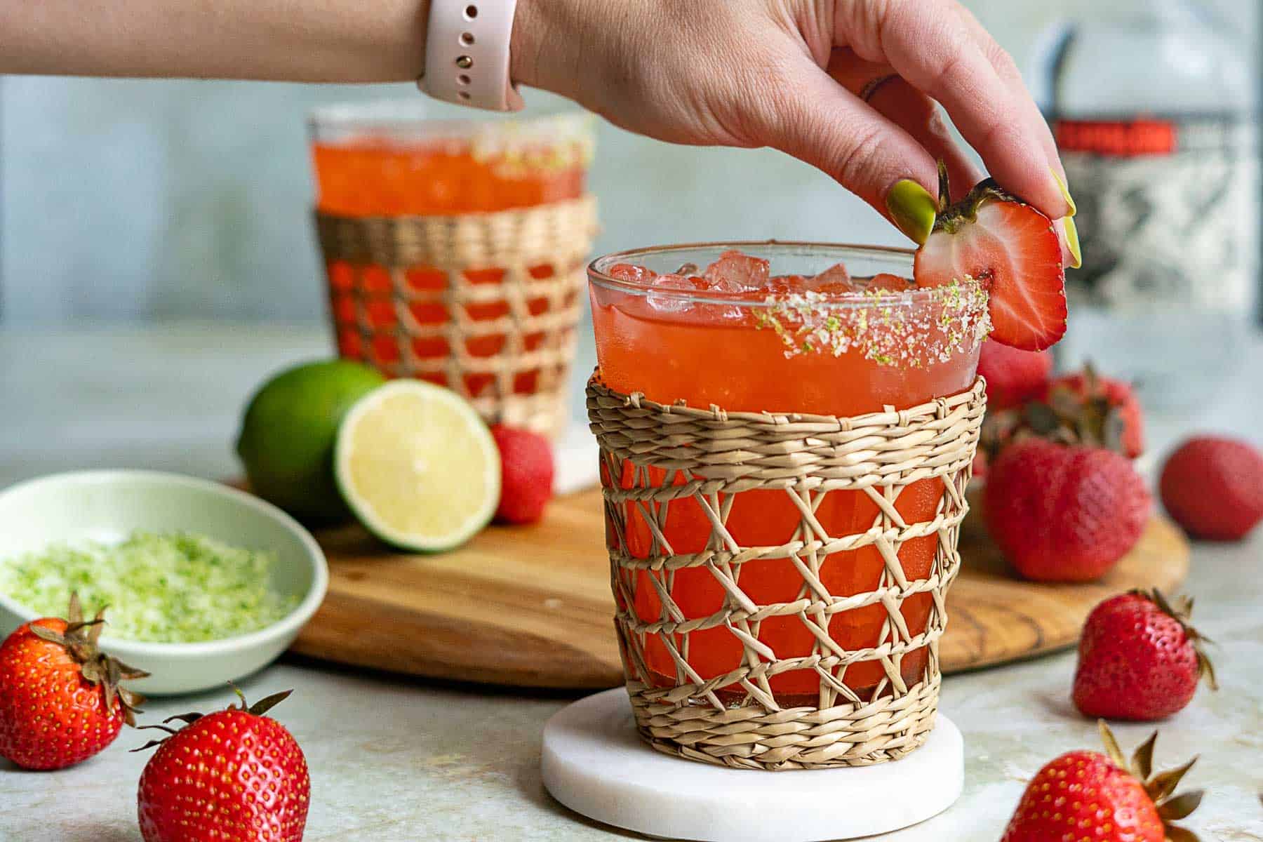 A hand garnishes a glass of strawberry margarita on the rocks with a fresh strawberry. The glass, in a wicker holder, sits on a marble coaster, surrounded by strawberries and a halved lime on a wooden board.