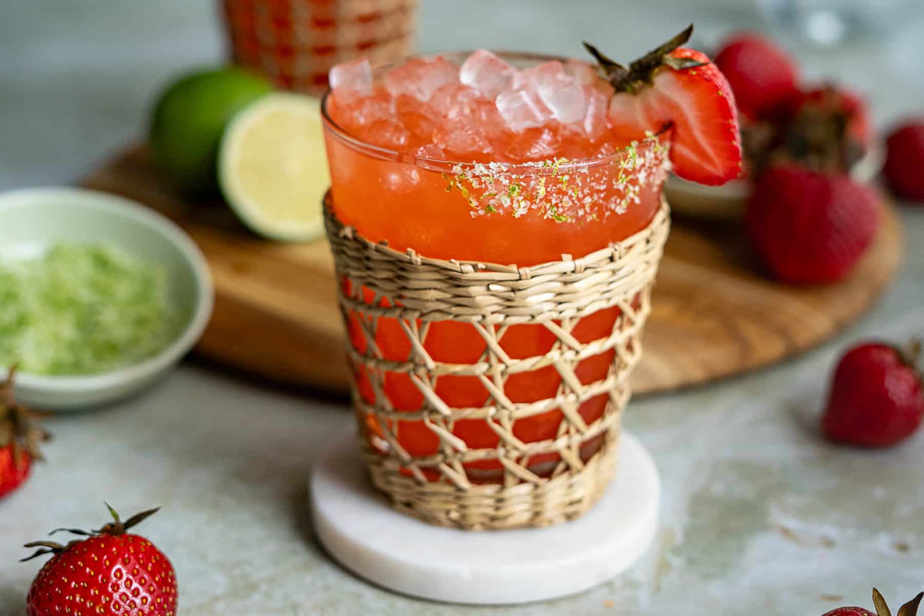A glass of strawberry margarita on the rocks with crushed ice, garnished with a fresh strawberry and sugared rim, sits in a woven holder on a coaster. Limes, a bowl of green sugar, and strawberries are in the background.