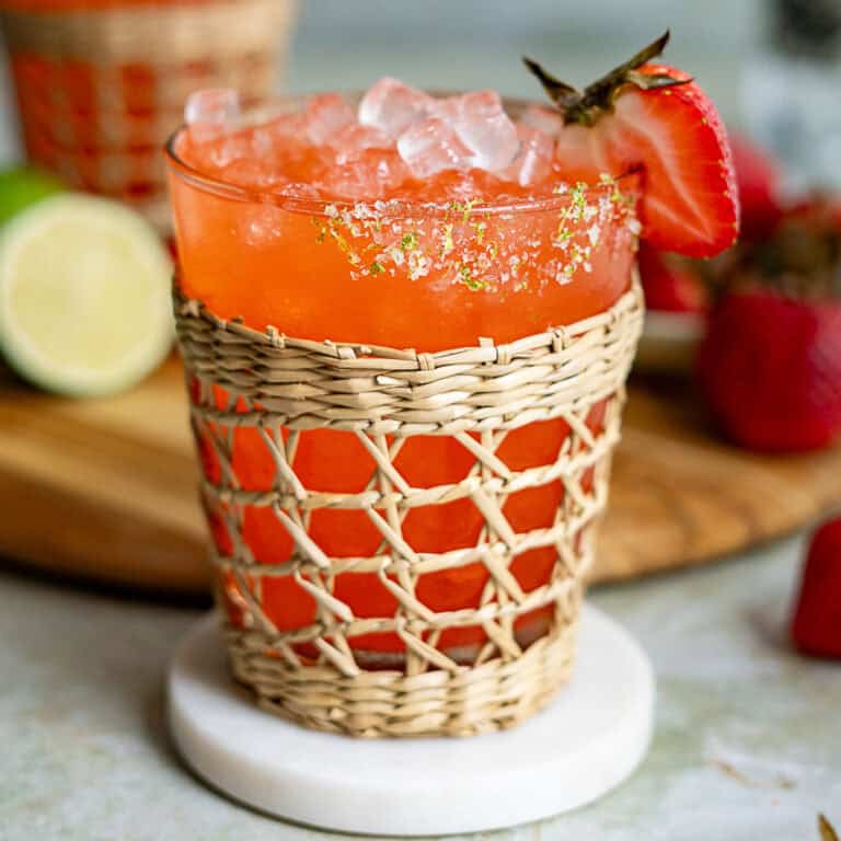 A vibrant red strawberry margarita cocktail with ice, garnished with a strawberry slice and lime zest, served in a glass wrapped with a decorative woven sleeve, placed on a white coaster with fresh strawberries and lime in the background.