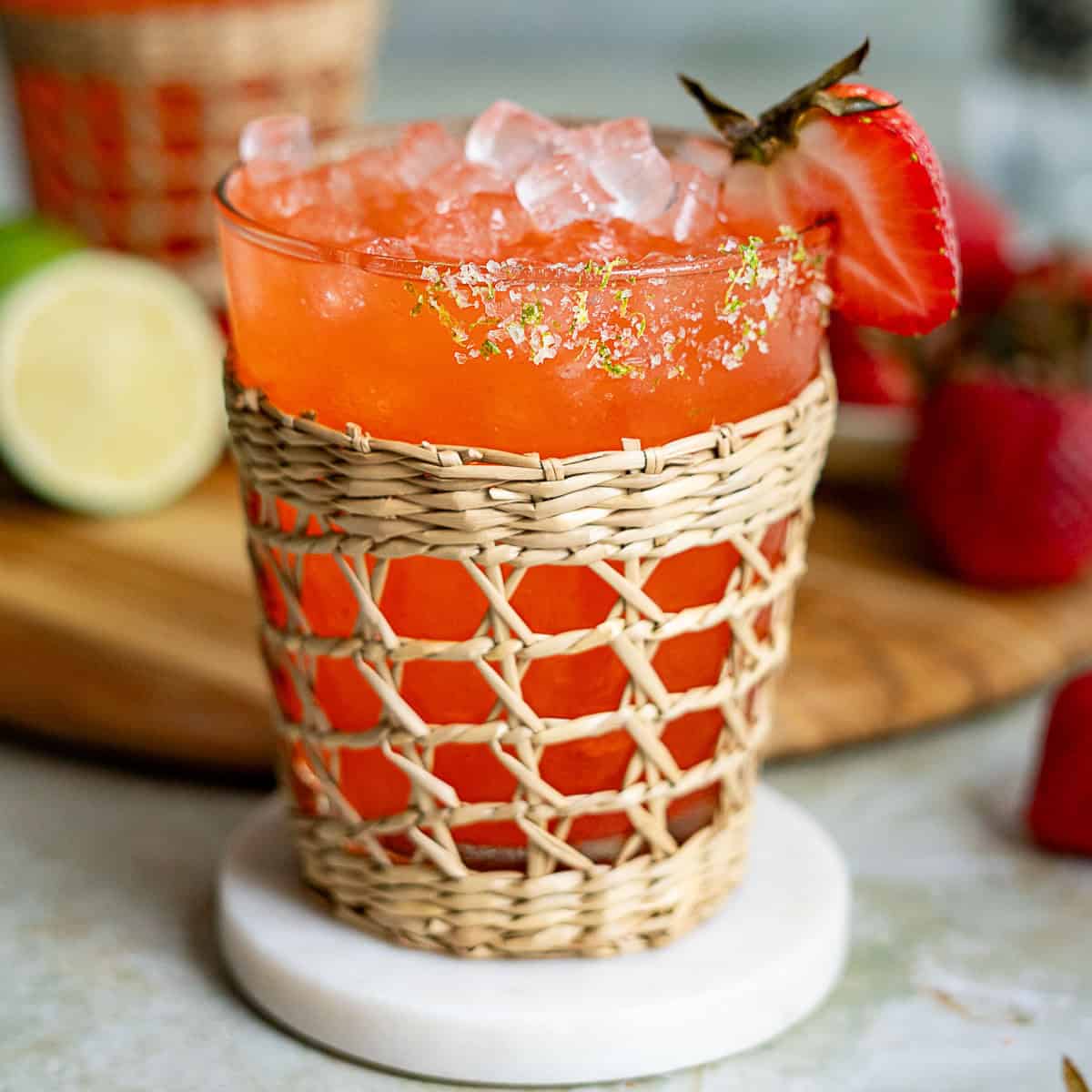 A vibrant red strawberry margarita cocktail with ice, garnished with a strawberry slice and lime zest, served in a glass wrapped with a decorative woven sleeve, placed on a white coaster with fresh strawberries and lime in the background.