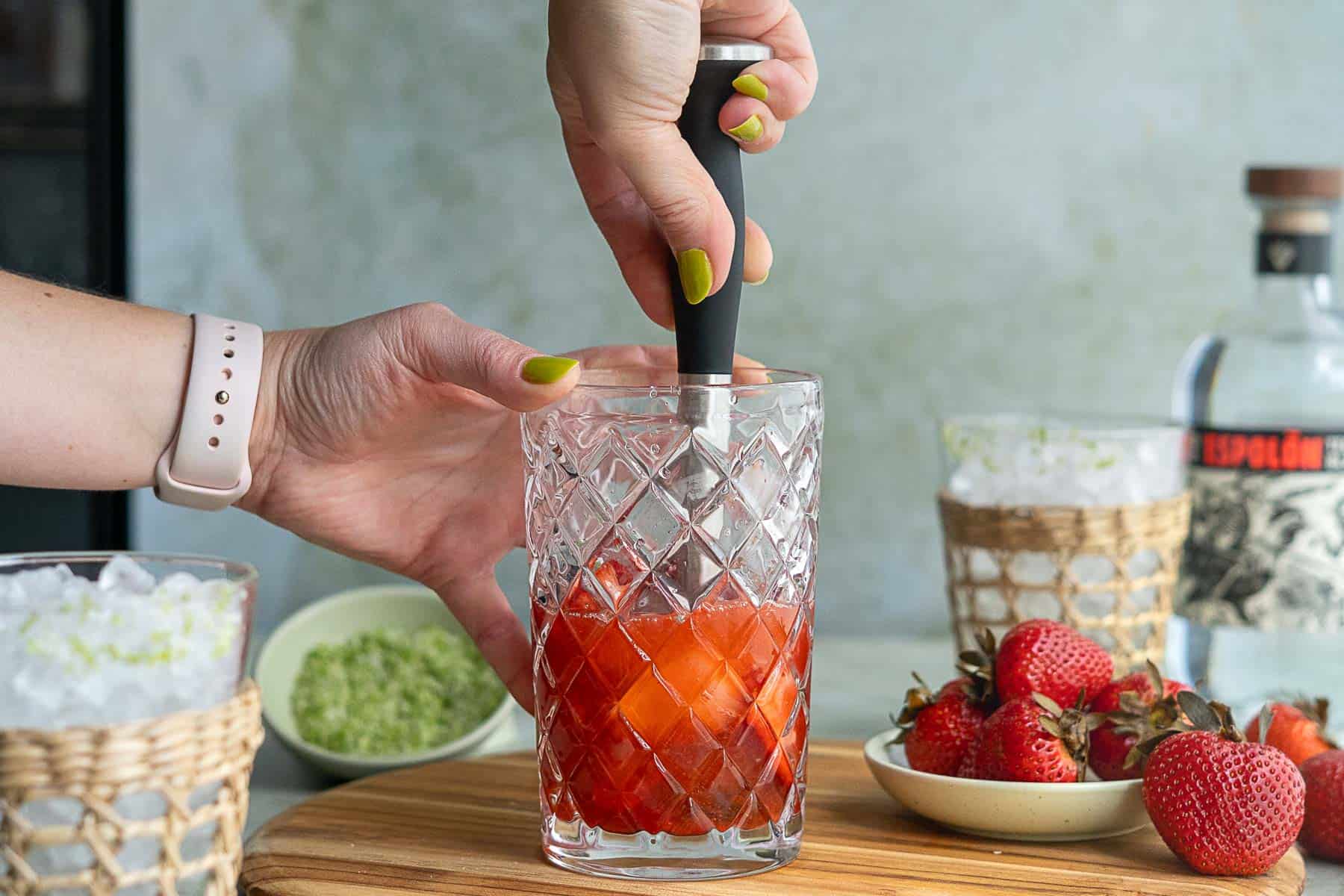 A hand uses a muddler to mash strawberries in a patterned glass, preparing a strawberry margarita on the rocks. Nearby are whole strawberries, green zest, and two glasses with ice. A bottle is blurred in the background.