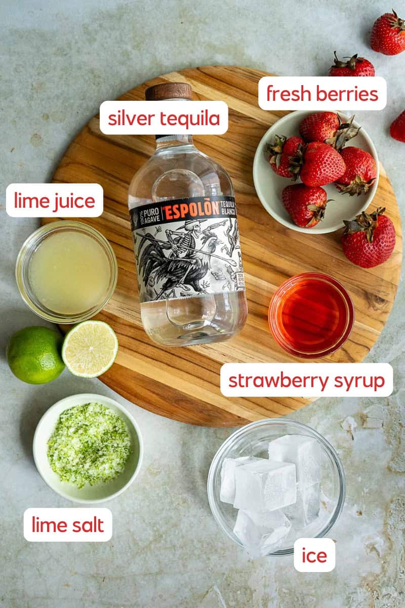 A round wooden board displays labeled ingredients for a strawberry margarita on the rocks: silver tequila, fresh berries, strawberry syrup, lime juice, lime salt, and a glass of ice cubes, all set on a light surface.