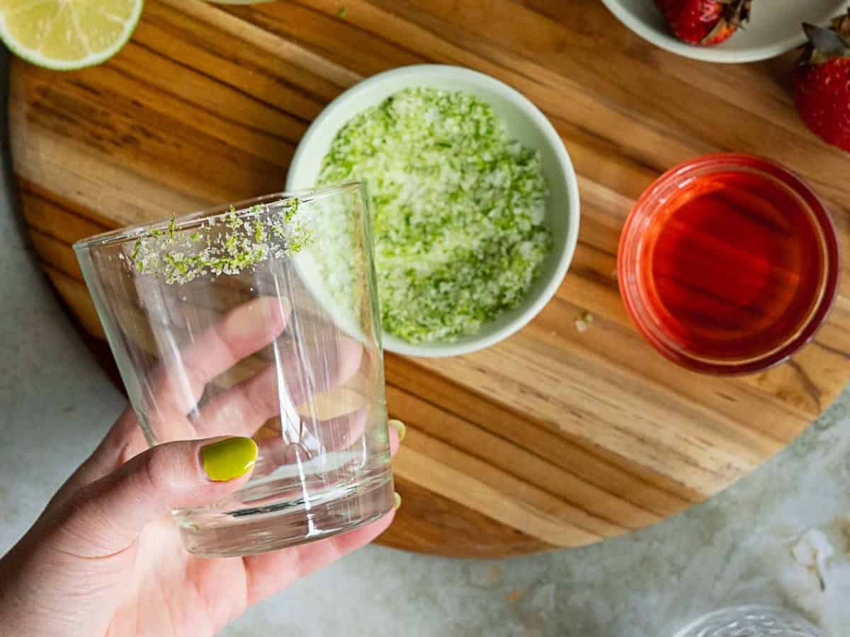 A hand with green-painted nails holds a strawberry margarita on the rocks, rimmed with lime and sugar, above a bowl of green sugar; nearby sit a red liquid in a glass, a lime wedge, and strawberries on a wooden board.