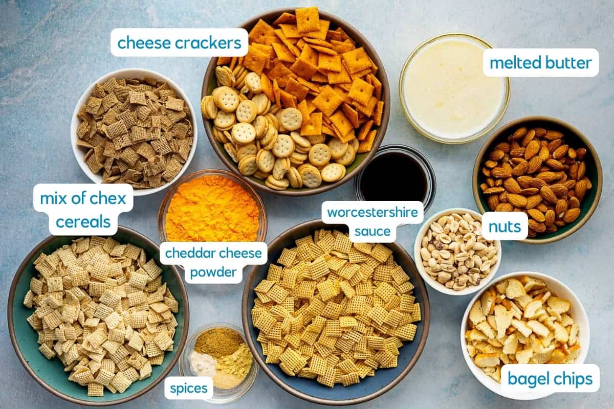 A flat lay of bowls containing ingredients for a cheddar Chex mix snack: cheese crackers, melted butter, mixed Chex cereals, cheddar cheese powder, Worcestershire sauce, nuts, bagel chips, and various spices—all labeled.