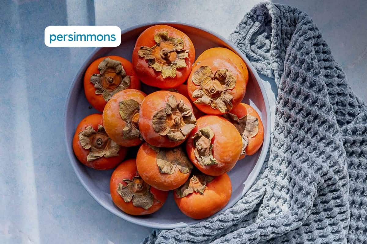 A bowl filled with ripe orange persimmons sits on a light blue surface next to a textured gray cloth. Sunlight casts shadows across the scene.