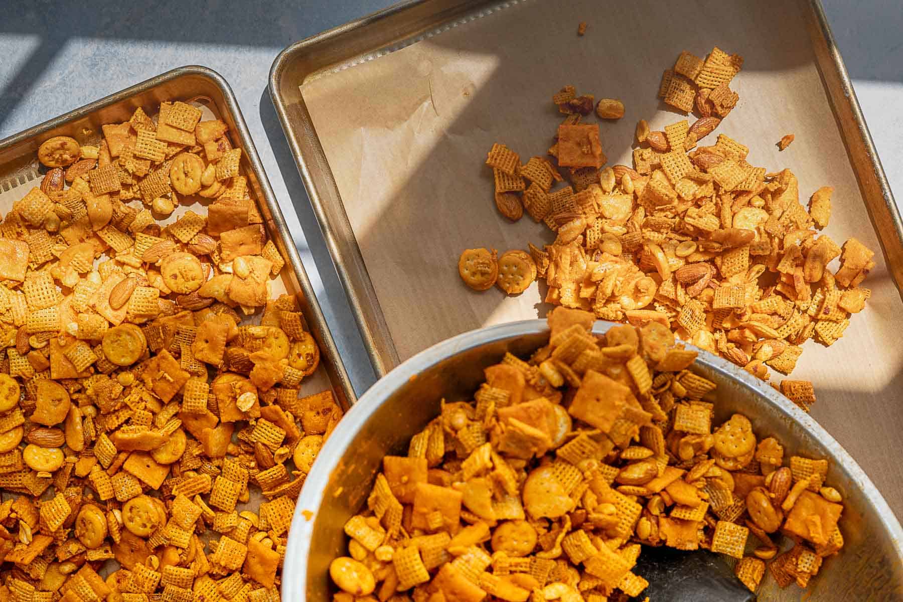 A close-up of homemade cheddar Chex mix with crackers, and cereal pieces spread on two baking sheets and in a large mixing bowl, viewed in natural sunlight.