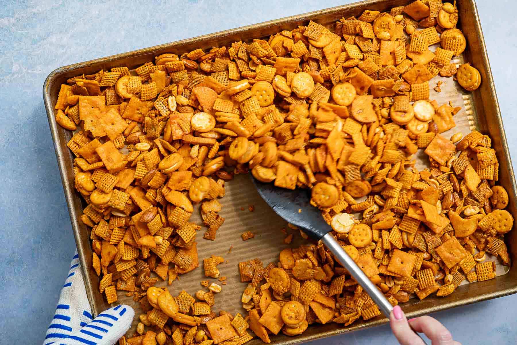 A hand uses a spatula to stir a cheddar Chex Mix snack blend of crackers, cereal, and nuts on a parchment-lined baking sheet. A gloved hand holds the tray steady.