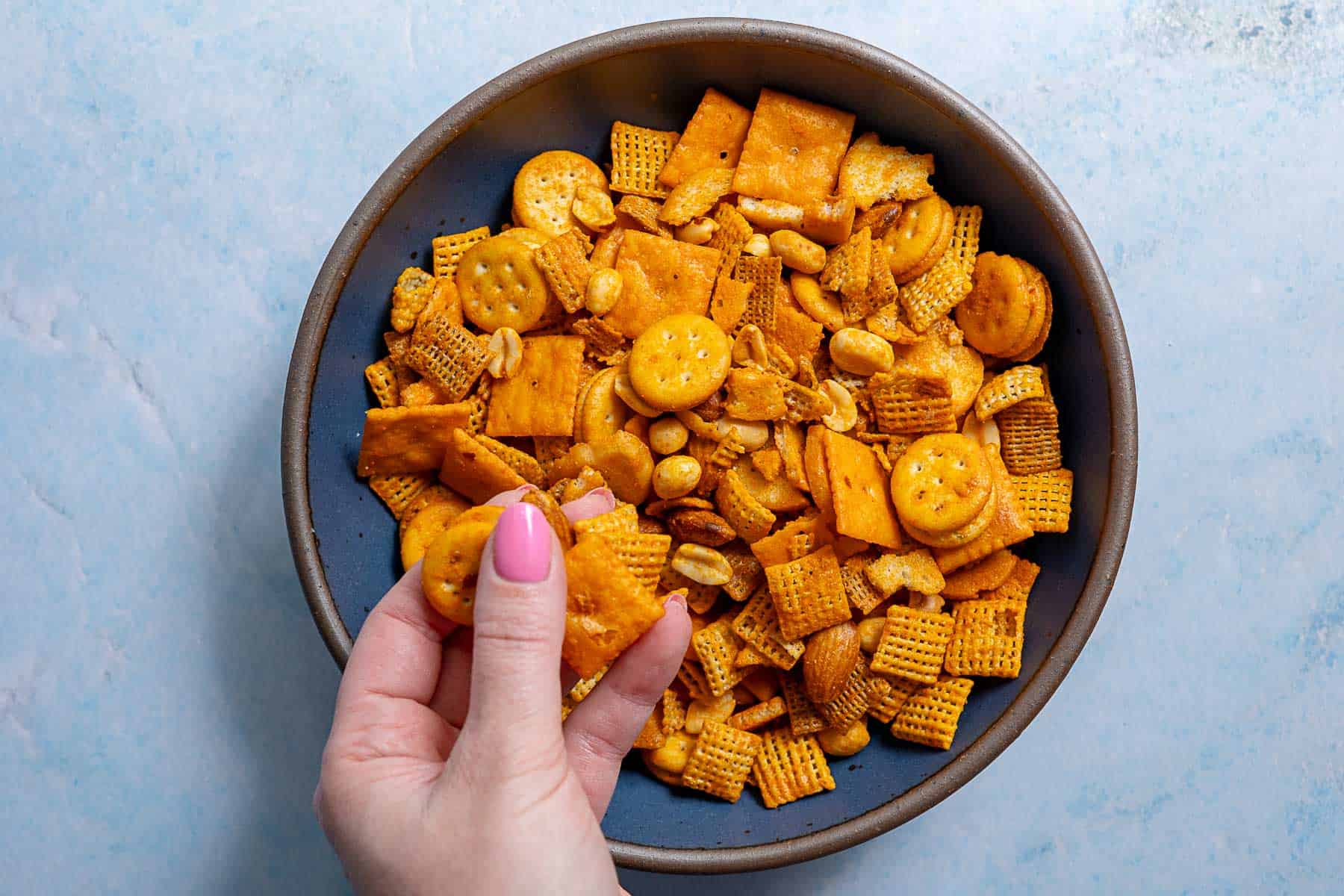 A hand picking up pieces of cheddar Chex Mix from a bowl filled with crackers, peanuts, and cereal on a light blue surface.