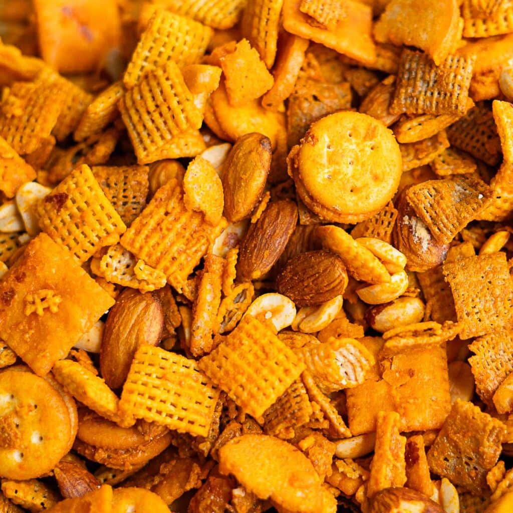 A close-up of cheddar Chex mix featuring square crackers, mini round crackers, pretzels, cereal squares, almonds, and peanuts, all coated in a savory orange seasoning.
