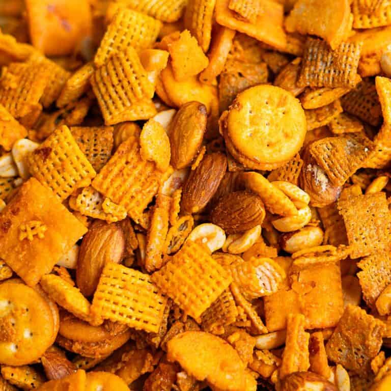 A close-up of cheddar Chex mix featuring square crackers, mini round crackers, pretzels, cereal squares, almonds, and peanuts, all coated in a savory orange seasoning.