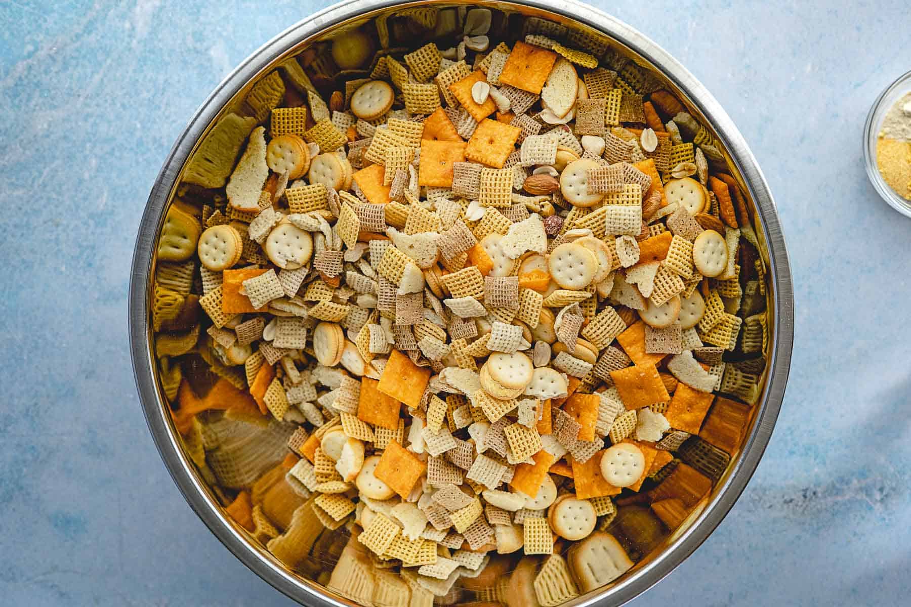 A large metal bowl filled with a cheddar Chex Mix snack blend of assorted crackers, Chex cereal, and peanuts, sitting on a light blue surface.