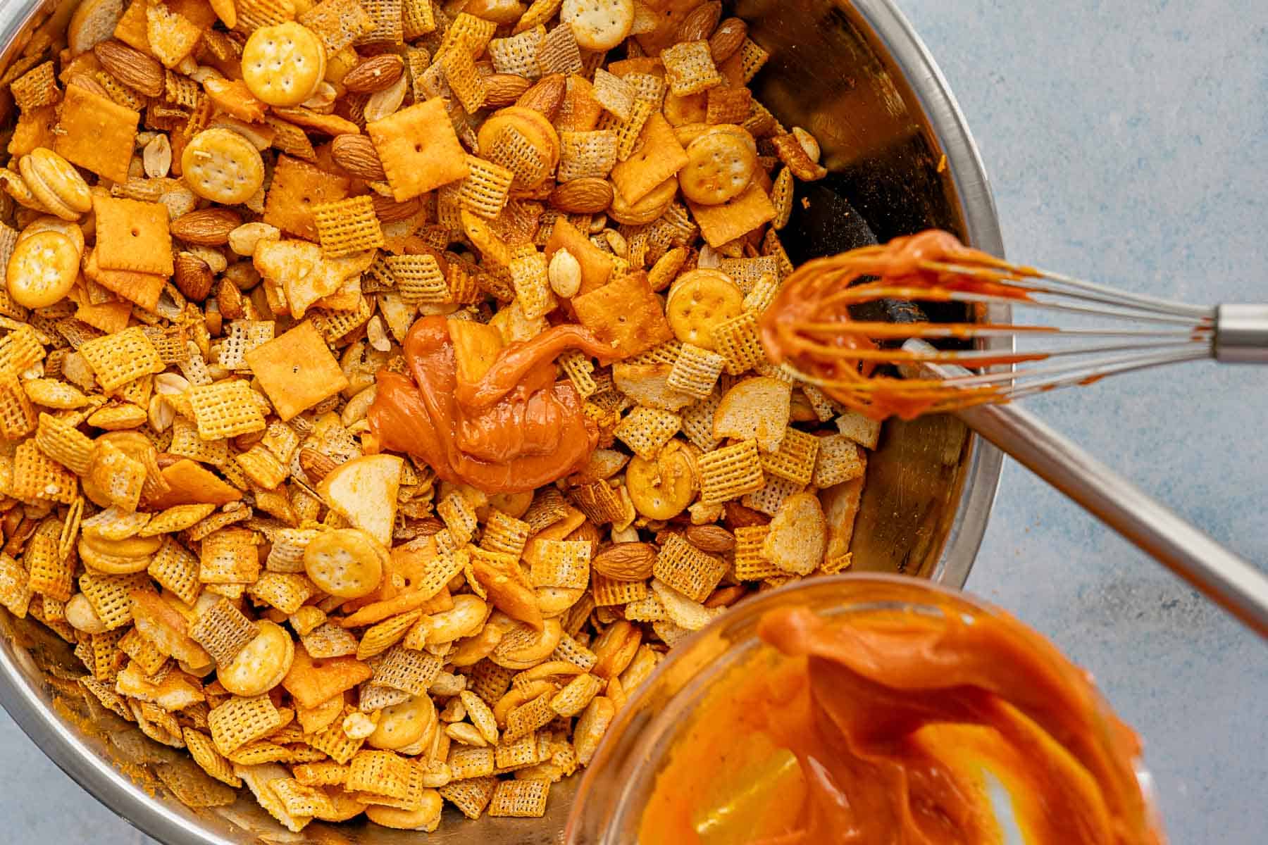 A mixing bowl filled with cheddar Chex mix—Chex cereal, mini crackers, almonds, and peanuts—being stirred with a whisk and a dollop of orange seasoning sauce. A glass bowl below holds extra sauce.