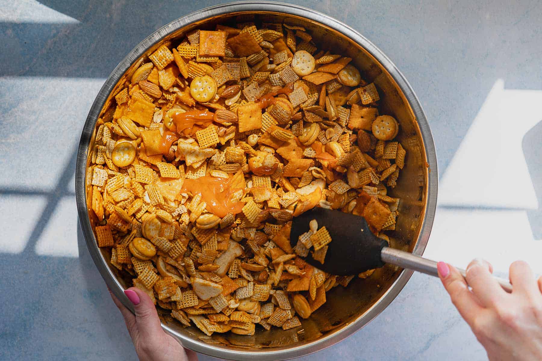 A person stirs a large metal bowl filled with homemade cheddar Chex mix—Chex cereal, crackers, and nuts—using a black spatula as sunlight casts shadows across the table.
