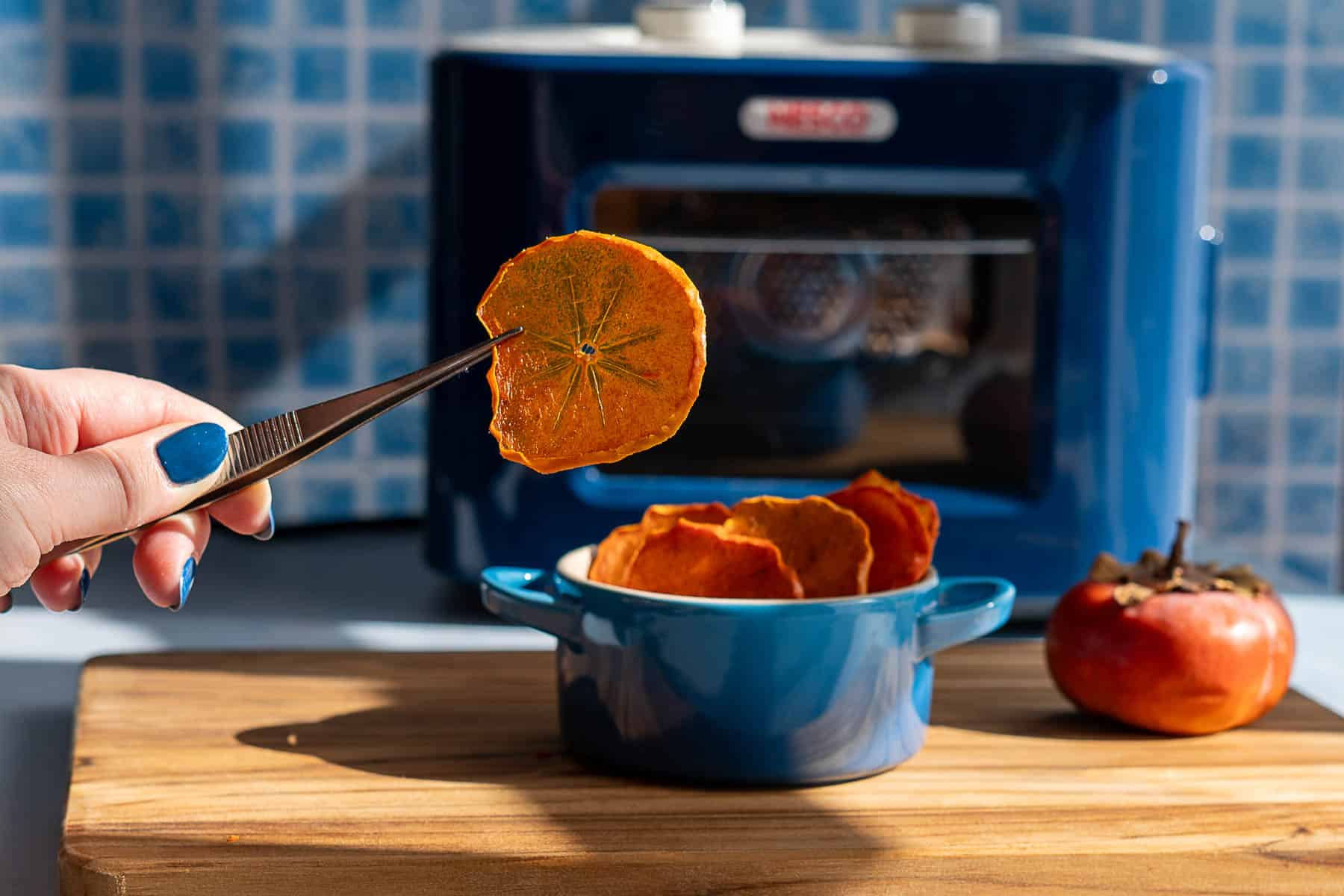 A hand holds a dehydrated persimmon slice with tweezers above a small blue bowl filled with more dehydrated persimmons, next to a whole persimmon and a blue kitchen appliance on a wooden surface.