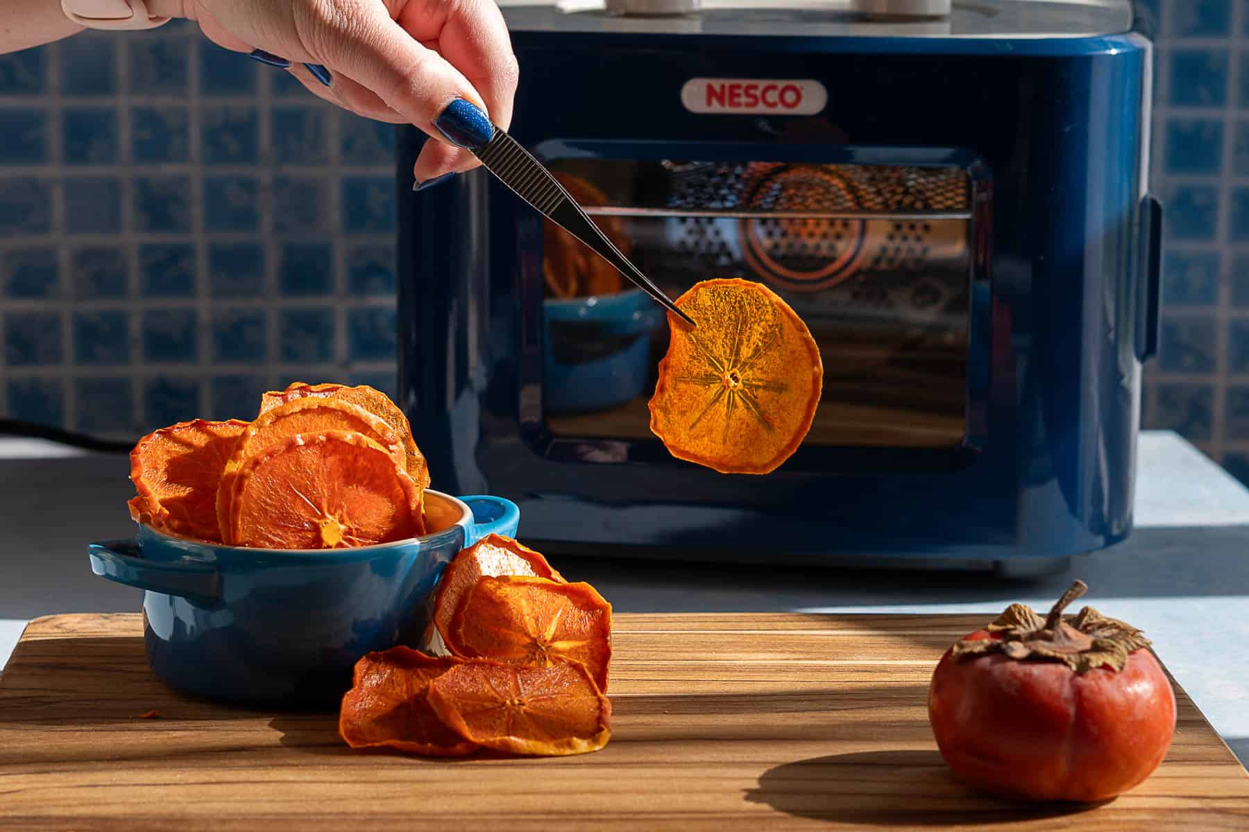 A hand uses tongs to hold a dehydrated persimmon slice above a bowl of dried persimmons on a wooden board. A fresh persimmon and a Nesco dehydrator are in the background.