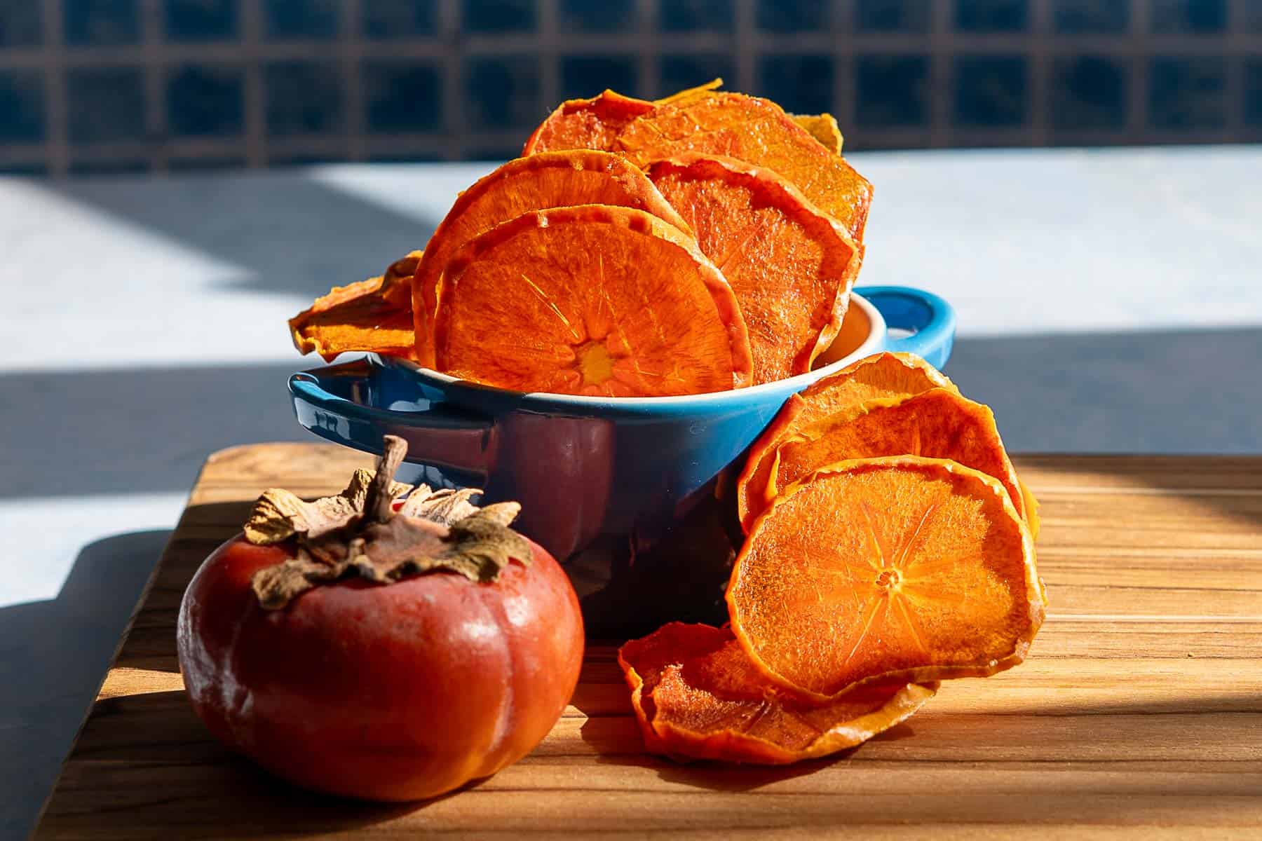 A blue bowl filled with dehydrated persimmons sits on a wooden board, with more dried slices scattered nearby and a whole fresh persimmon placed beside the bowl.