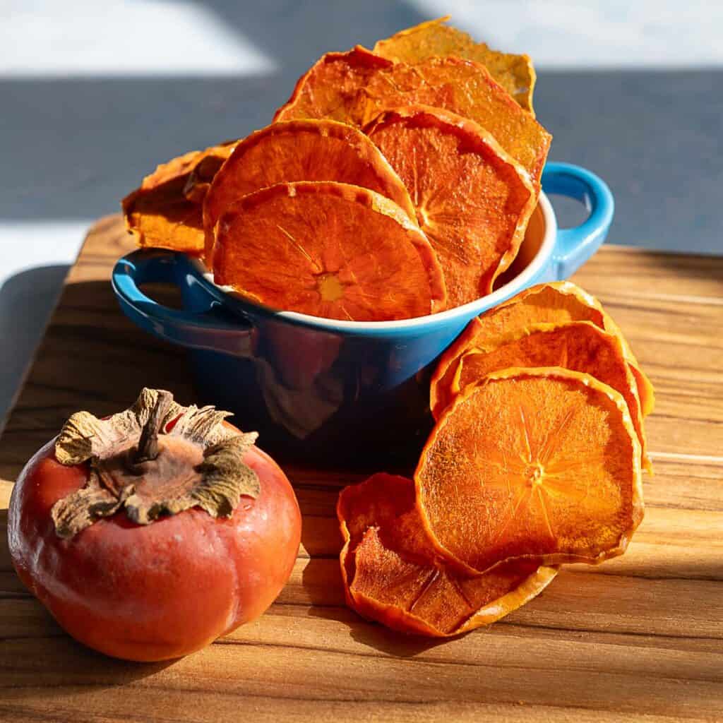 A blue bowl filled with dehydrated persimmons sits on a wooden board, with more dried slices scattered nearby and a whole fresh persimmon placed beside the bowl.