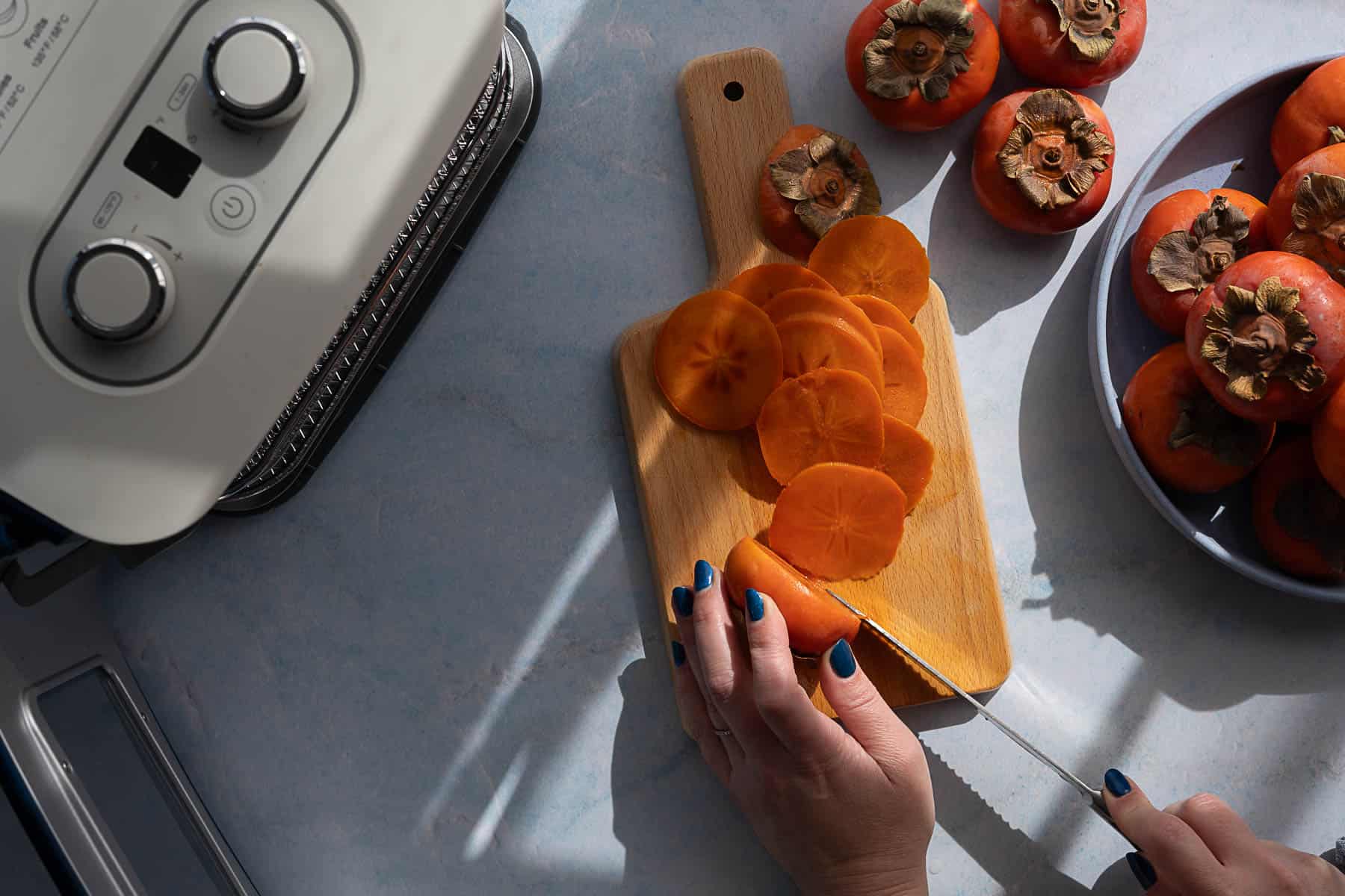 A person with blue nail polish slices a persimmon on a wooden cutting board. Several whole persimmons are nearby, and a kitchen appliance is visible on the counter.