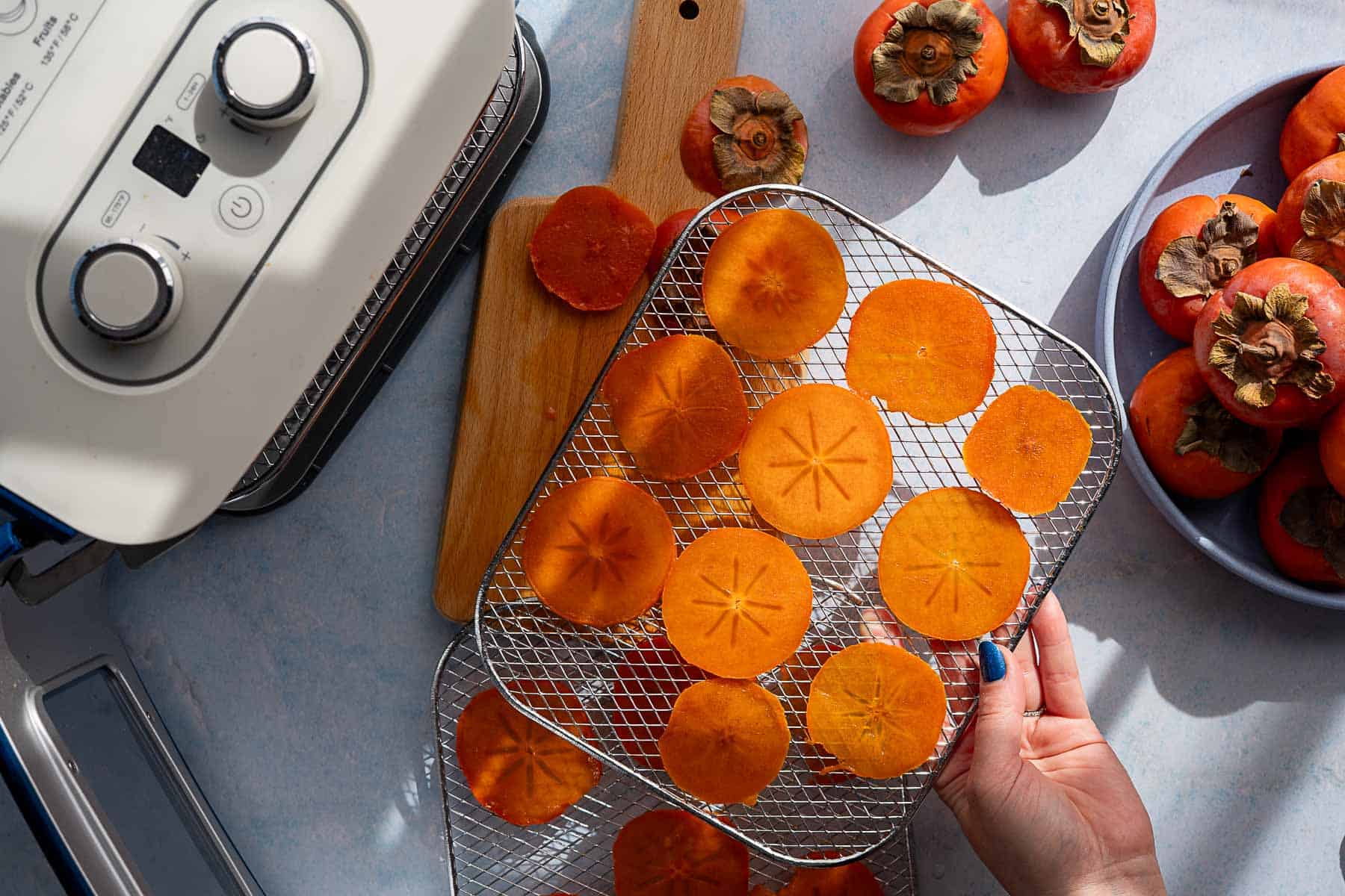 Sliced persimmons arranged on a dehydrator rack, with whole persimmons nearby on a plate and cutting board; a hand holds the rack next to an air fryer, ready to make delicious dehydrated persimmons on a light-colored countertop.