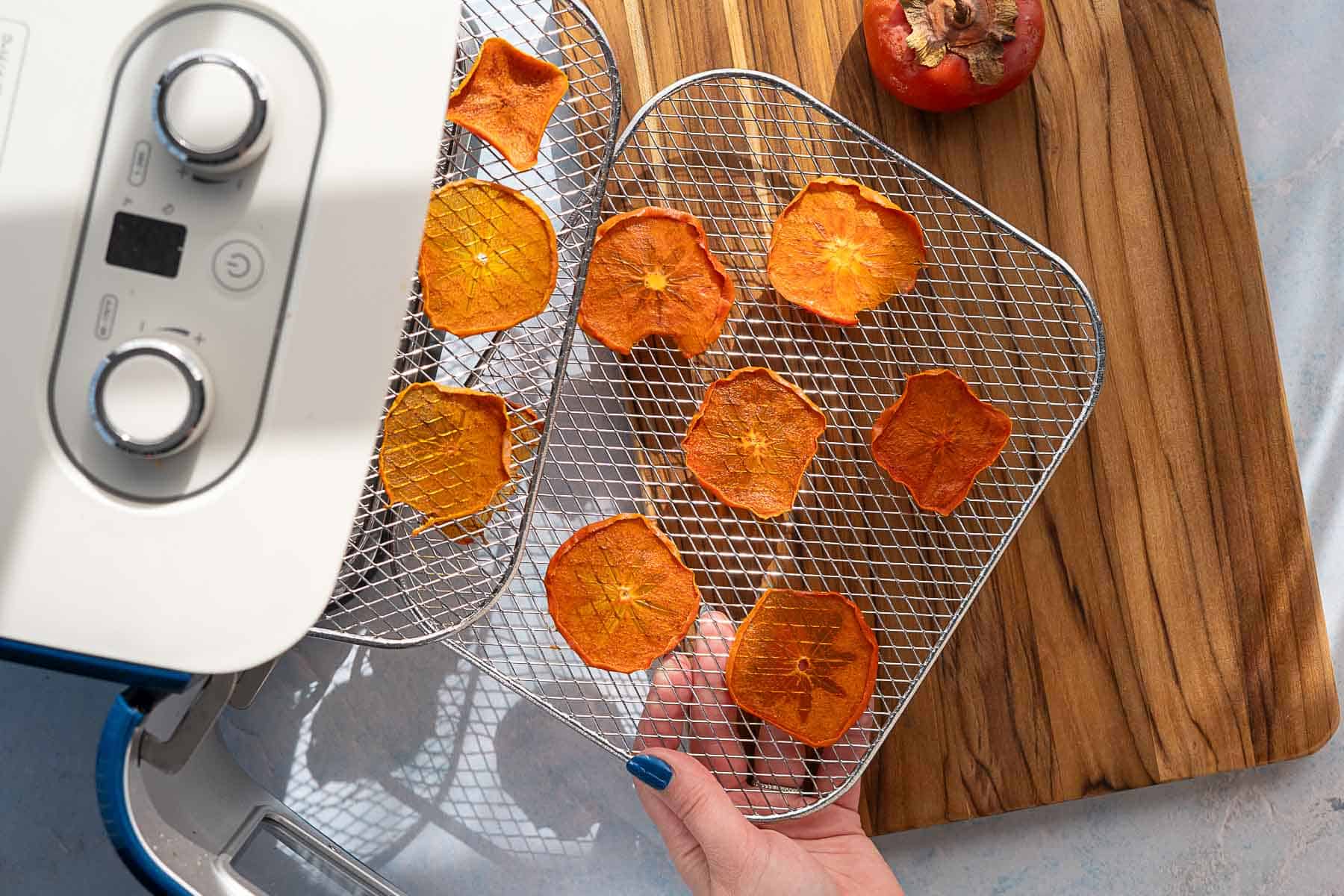 A hand holds a dehydrator tray with dehydrated persimmons on a wire rack, next to a whole persimmon resting on a wooden cutting board. These persimmons were sliced thinner and dehydrated at a lower temperature for a longer period of time.