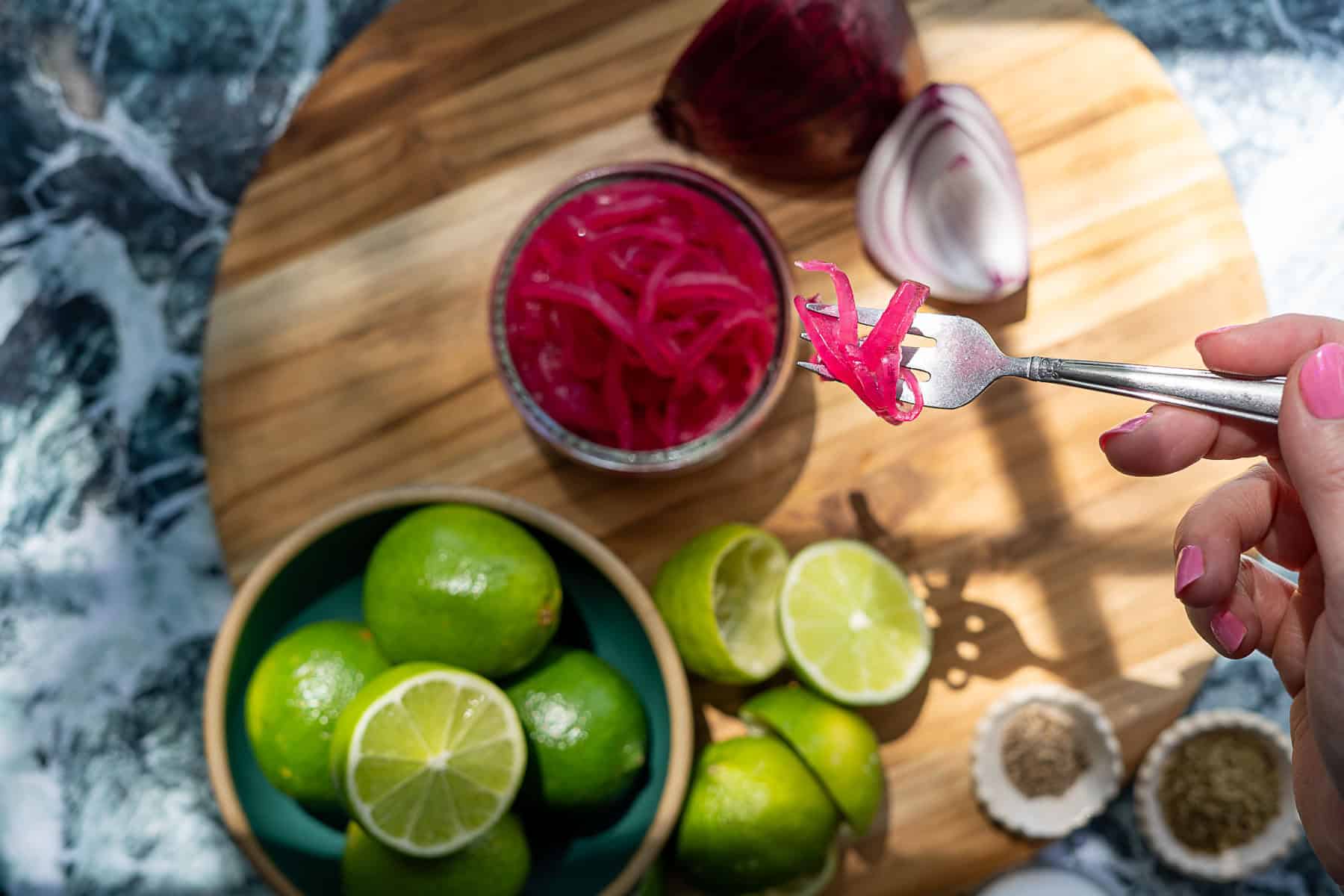 A hand with pink nails holds a fork with pickled red onions above a wooden board displaying a jar of pickled red onions, a bowl of limes, lime slices, a red onion, and small bowls of spices.