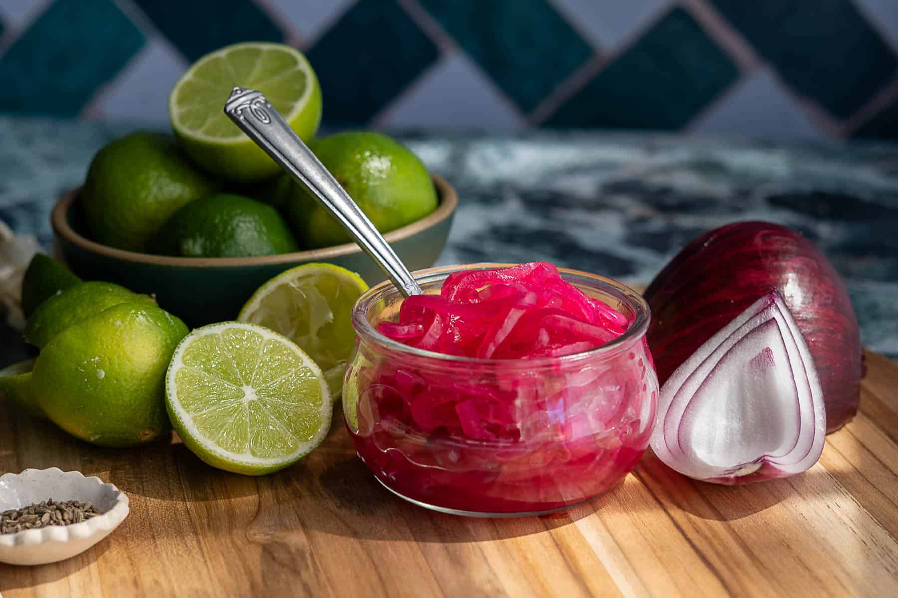 A small glass bowl of pickled red onions with a spoon sits on a wooden surface, surrounded by fresh limes, lime halves, spiced seeds, and a halved red onion. In the background, a bowl of whole limes complements the vibrant pickled red onions.