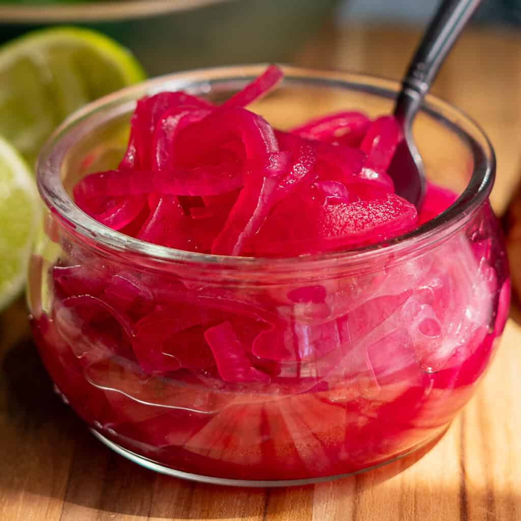A small glass bowl of pickled red onions with a spoon sits on a wooden surface, surrounded by fresh limes, lime halves, spiced seeds, and a halved red onion. In the background, a bowl of whole limes complements the vibrant pickled red onions.