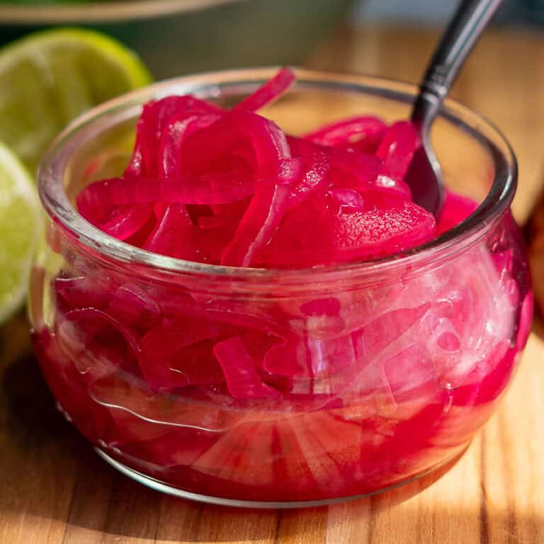 A small glass bowl of pickled red onions with a spoon sits on a wooden surface, surrounded by fresh limes, lime halves, spiced seeds, and a halved red onion. In the background, a bowl of whole limes complements the vibrant pickled red onions.