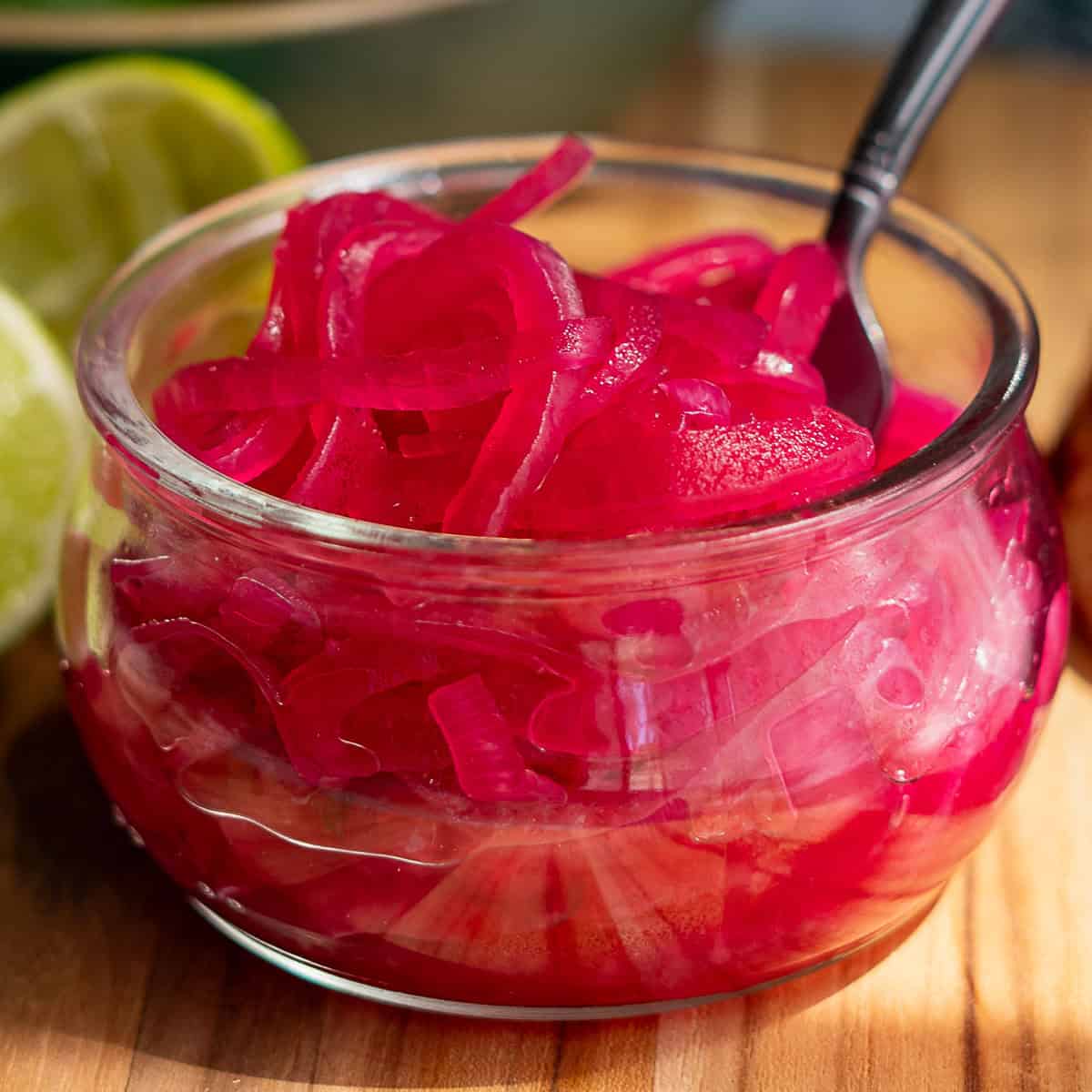 A small glass bowl of pickled red onions with a spoon sits on a wooden surface, surrounded by fresh limes, lime halves, spiced seeds, and a halved red onion. In the background, a bowl of whole limes complements the vibrant pickled red onions.