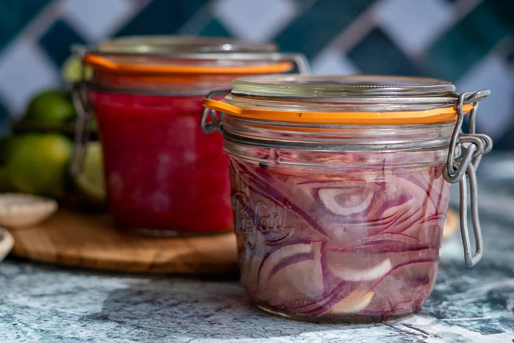 Two glass jars with orange rubber seals sit on a countertop—one filled with pickled red onions in liquid, the other with a more vibrant batch of red onions that has marinated overnight—surrounded by limes and set against a blurred tile background.