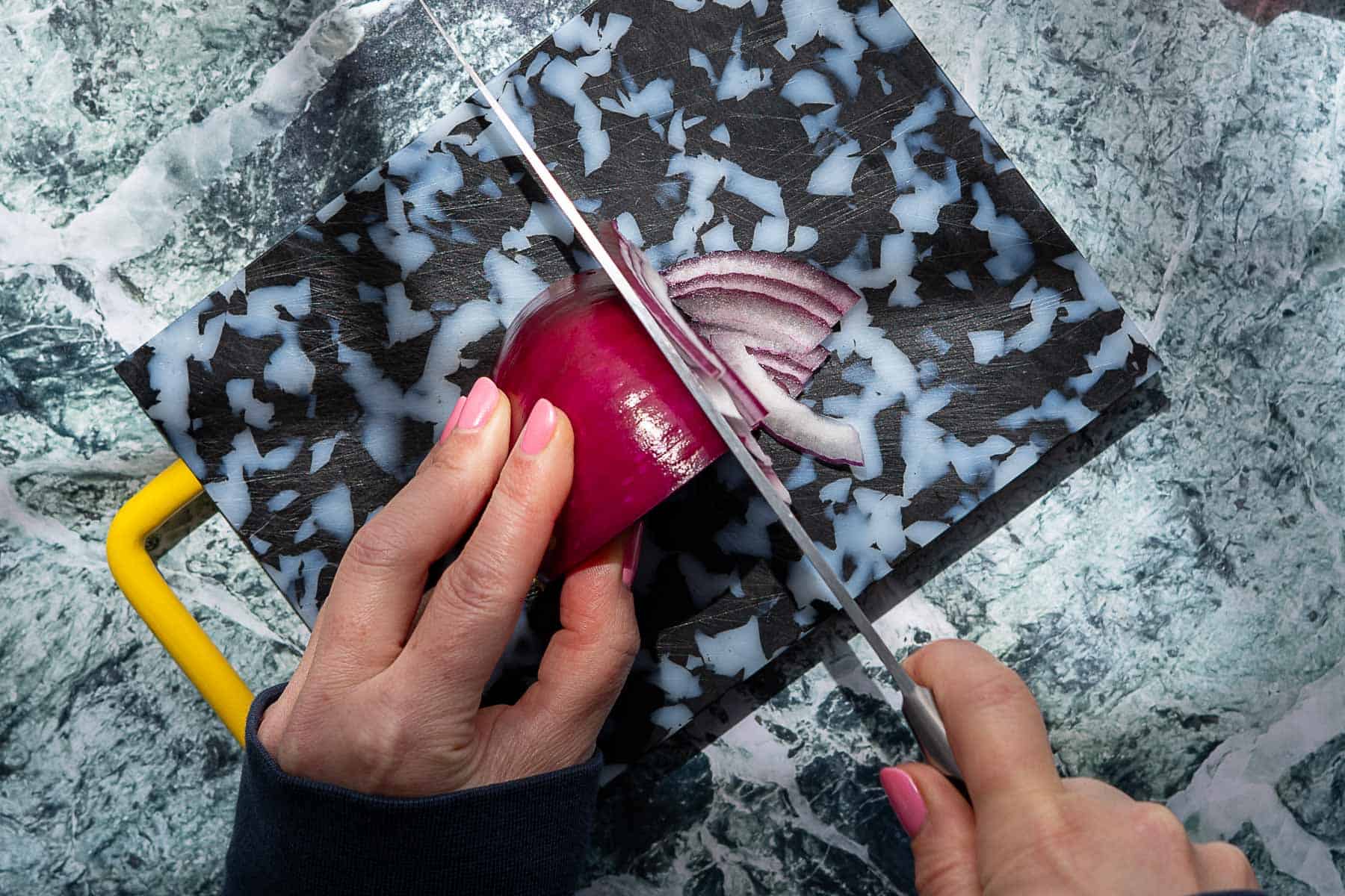 A person with pink nail polish slices a red onion on a black and white patterned cutting board with a yellow handle, preparing it for pickled red onions.