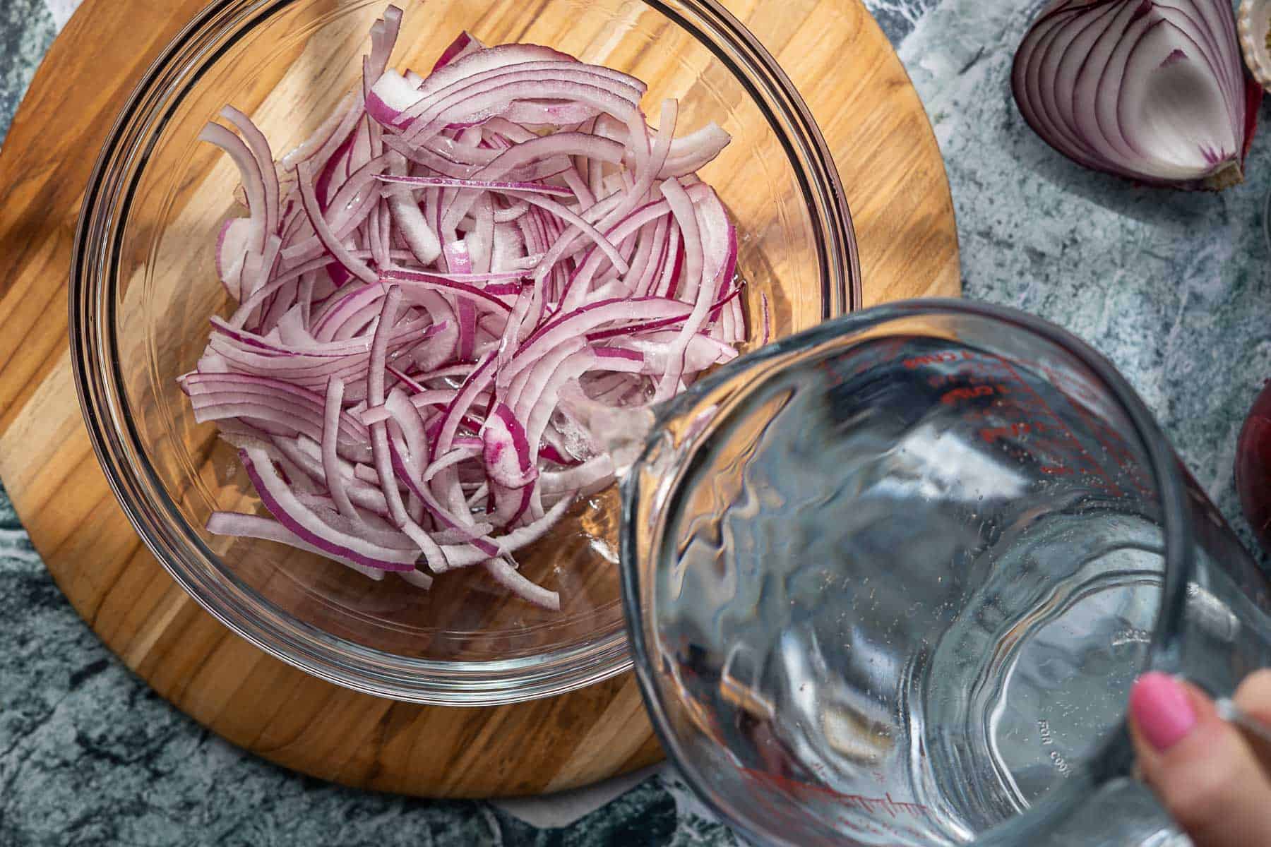 A clear bowl with thinly sliced red onions sits on a wooden board. A hand pours water from a glass pitcher into the bowl, starting the process of making pickled red onions. Nearby, a cut red onion rests on a gray surface.