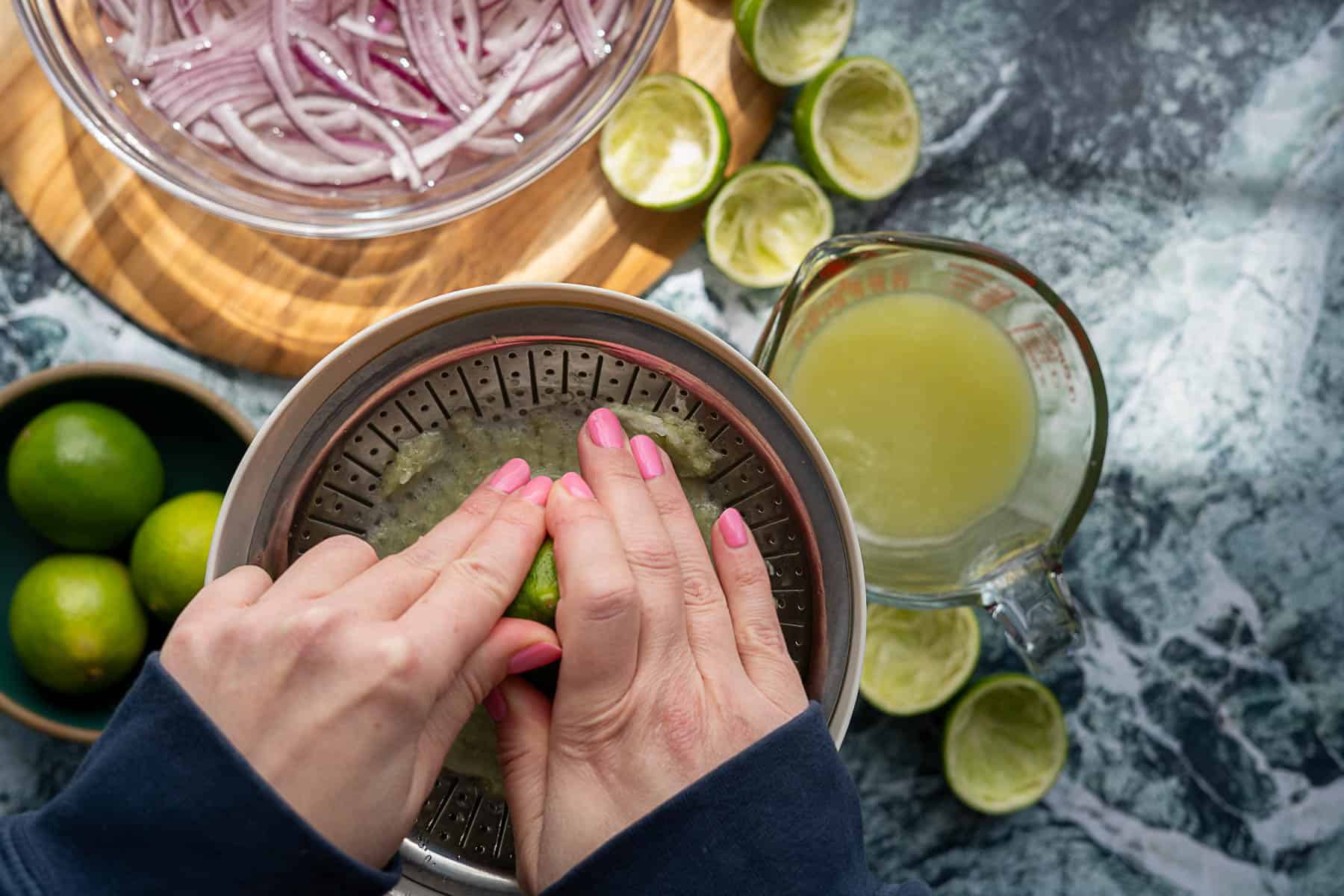 Hands with pink nail polish squeeze a lime on a juicer. Nearby are a bowl of sliced pickled red onions, whole and halved limes, and a measuring cup filled with lime juice on a marble countertop.