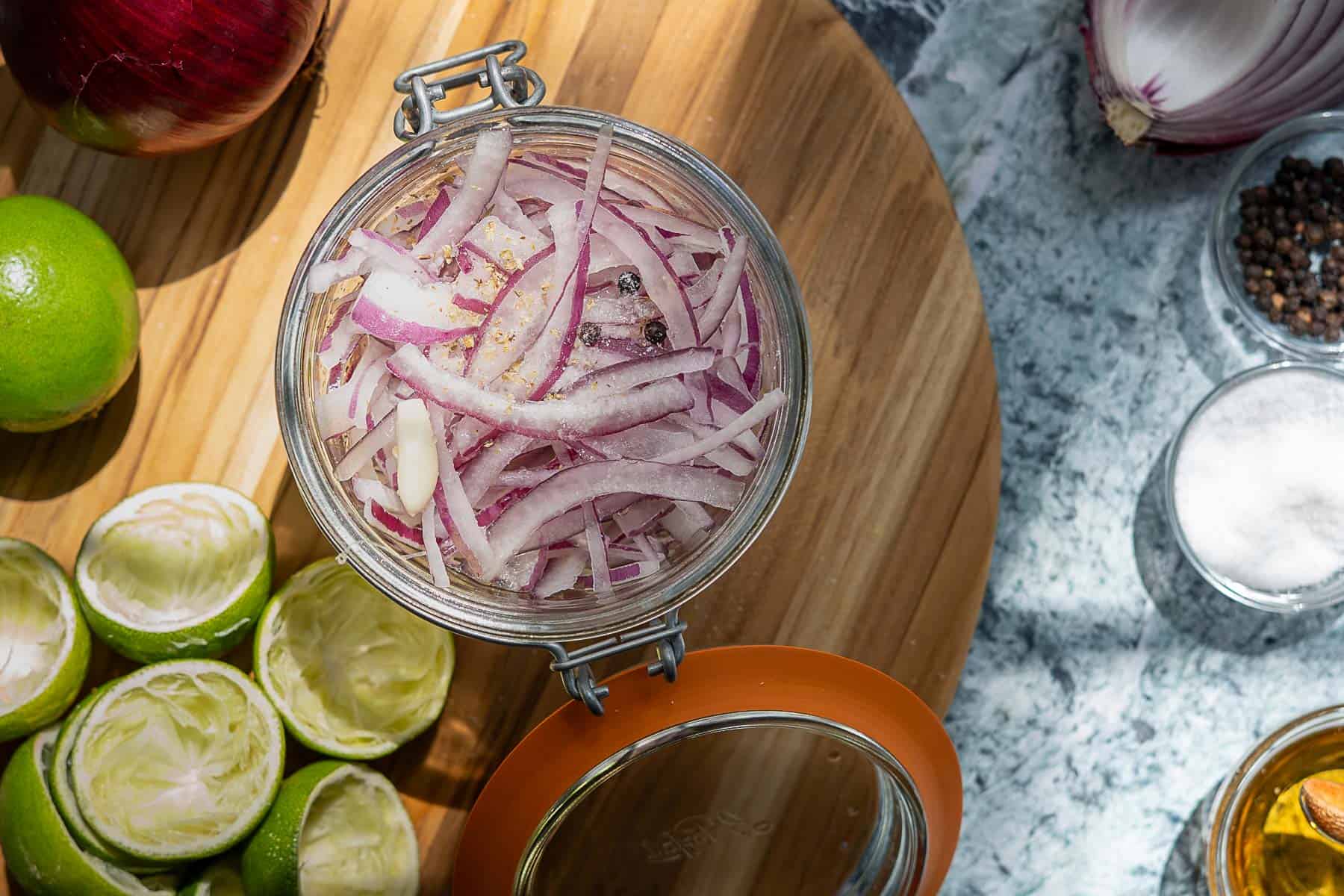 A glass jar of pickled red onions, packed with spices and brine, sits on a wooden surface. Surrounded by squeezed lime halves, a red onion, a bowl of salt, peppercorns, and a small bottle of oil.