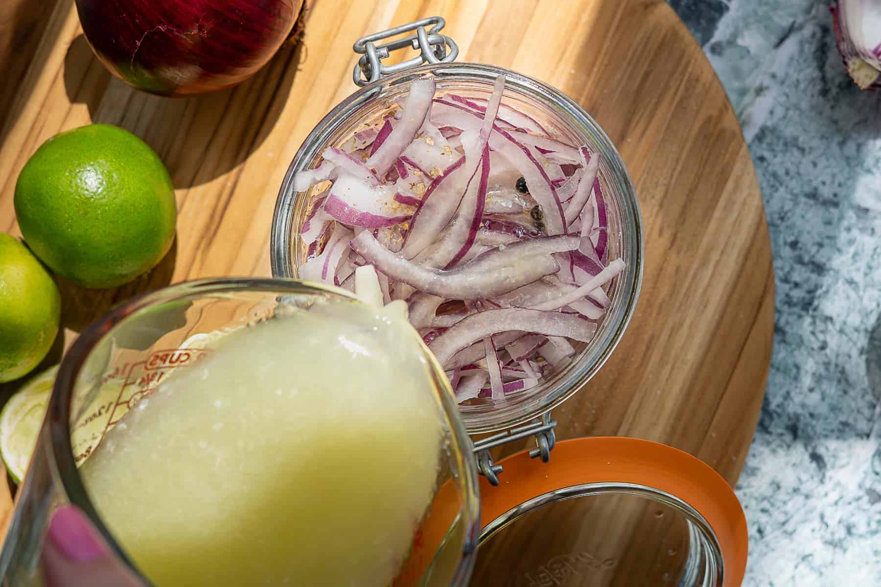 A jar of pickled red onions is being topped with freshly squeezed lime juice. Whole limes, a red onion, and a wooden cutting board are visible in the background.