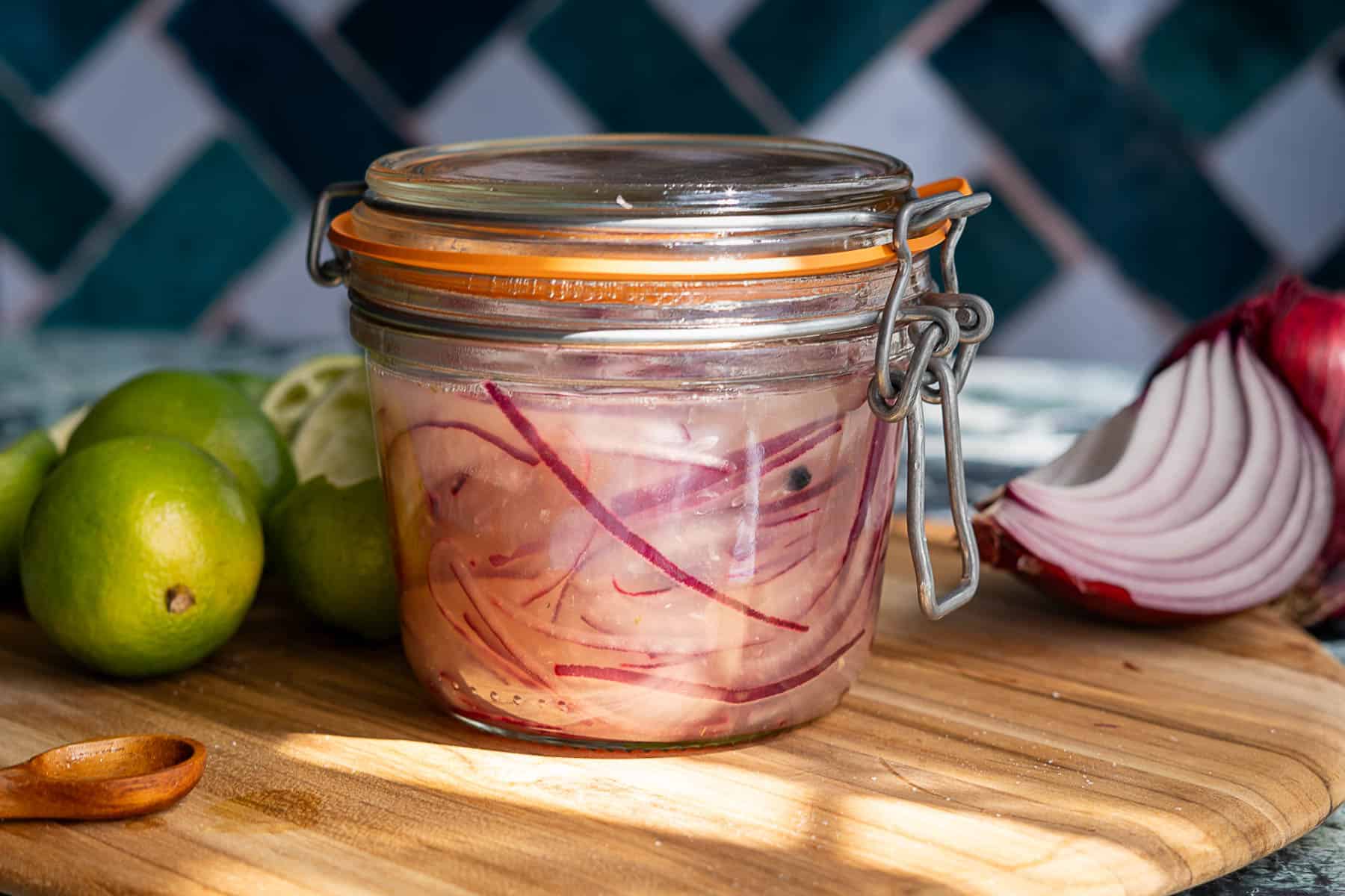 A glass jar of pickled red onions sits on a wooden cutting board, surrounded by whole limes and a sliced red onion. The background features a blue and white tiled wall, highlighting the vibrant color of the pickled red onions.