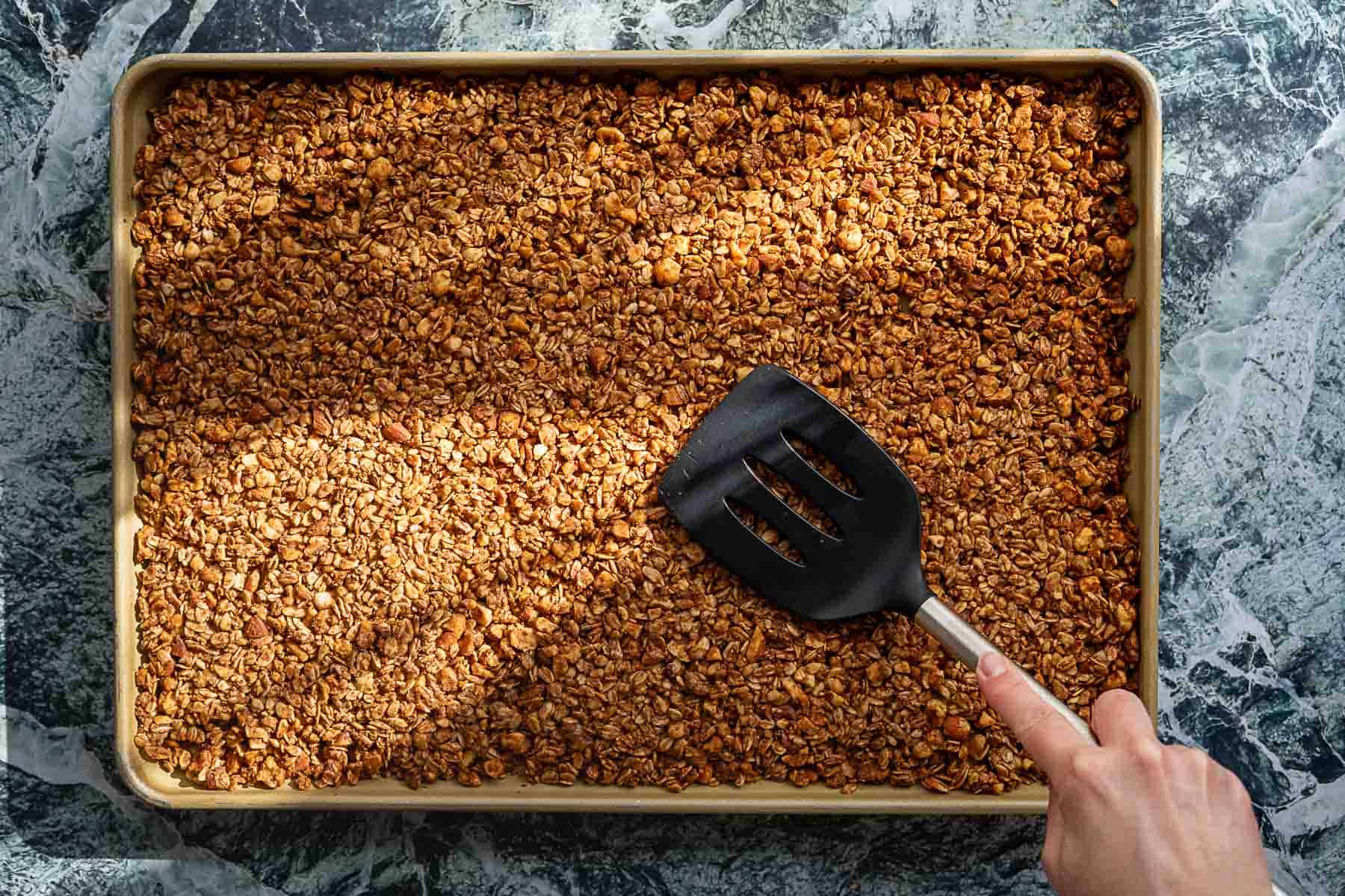 A hand holding a black spatula is stirring golden-brown sourdough discard granola on a baking sheet, set on a marbled countertop.