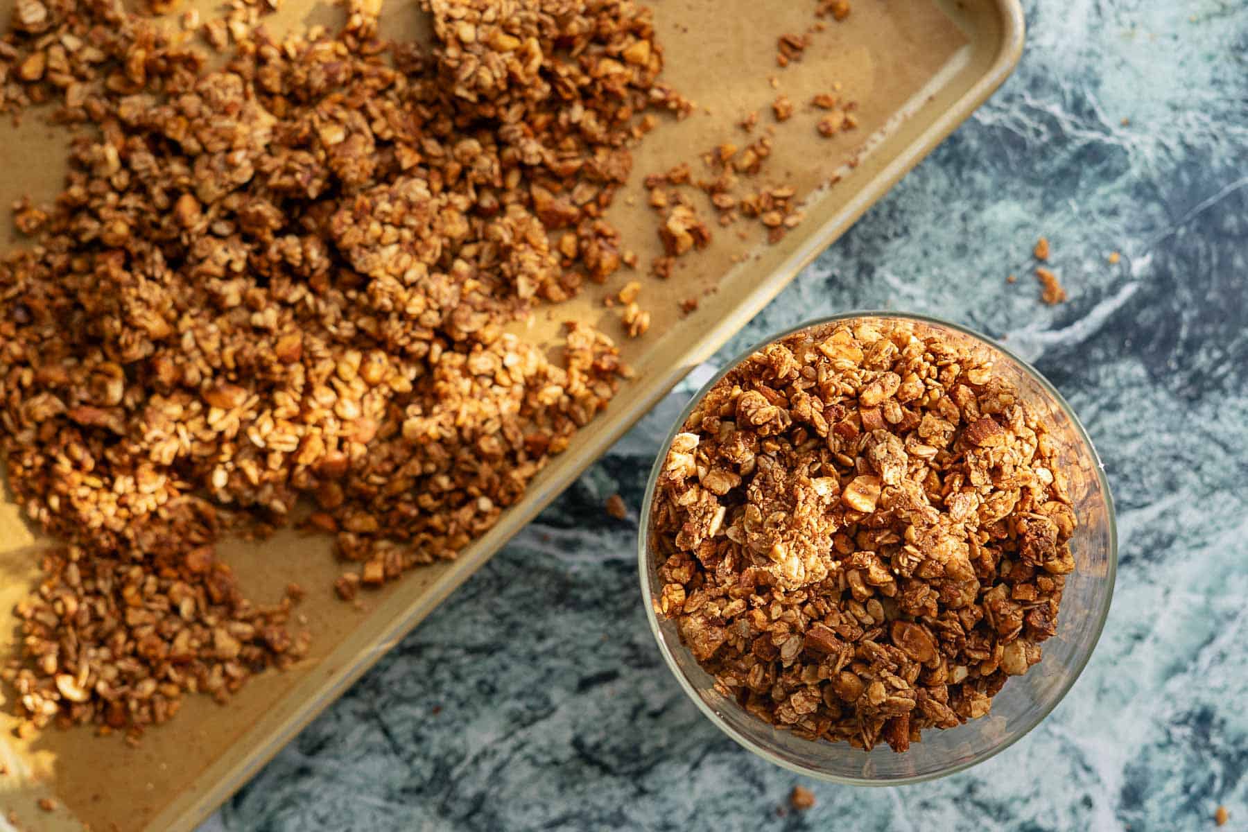 A glass filled with homemade sourdough discard granola sits on a marble surface next to a baking sheet covered with more golden brown, chunky granola.