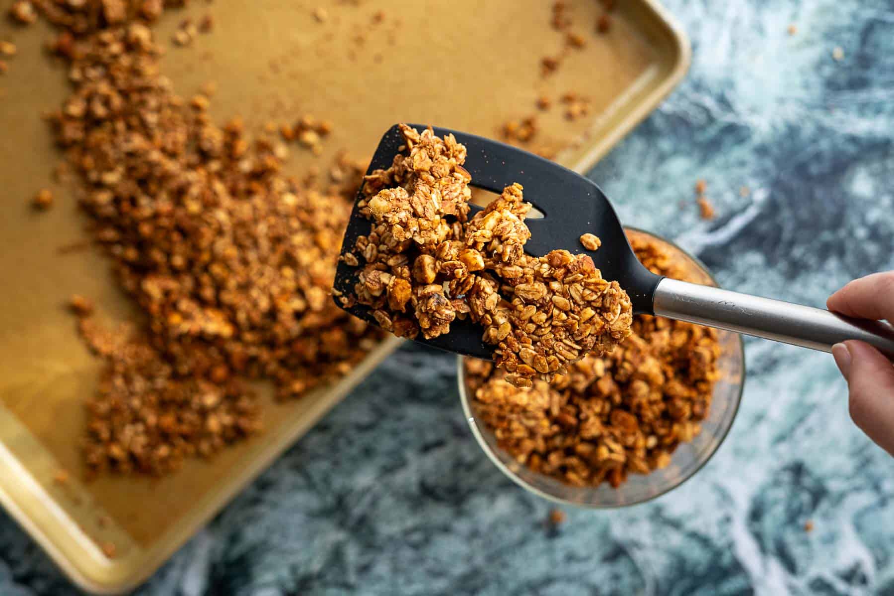 A hand uses a black spatula to scoop homemade sourdough discard granola from a bowl, with a baking sheet of scattered granola on a marble counter in the background.