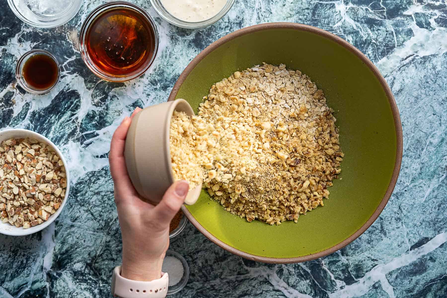 A hand pours chopped nuts into a green bowl filled with oats and sourdough discard granola on a marble countertop, surrounded by small bowls containing ingredients like nuts, syrup, and vanilla extract.
