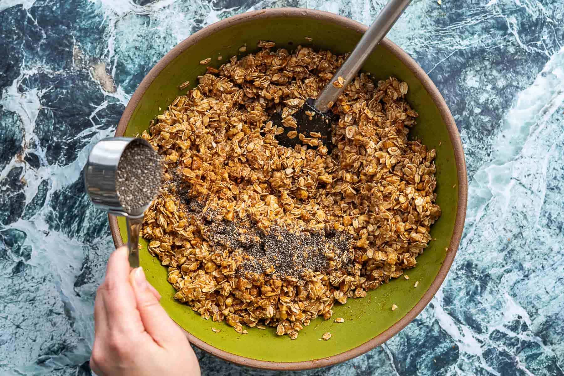 A hand pours chia seeds from a metal measuring cup into a green bowl filled with oats, sourdough discard granola, and other ingredients, being stirred with a spatula on a marble countertop.