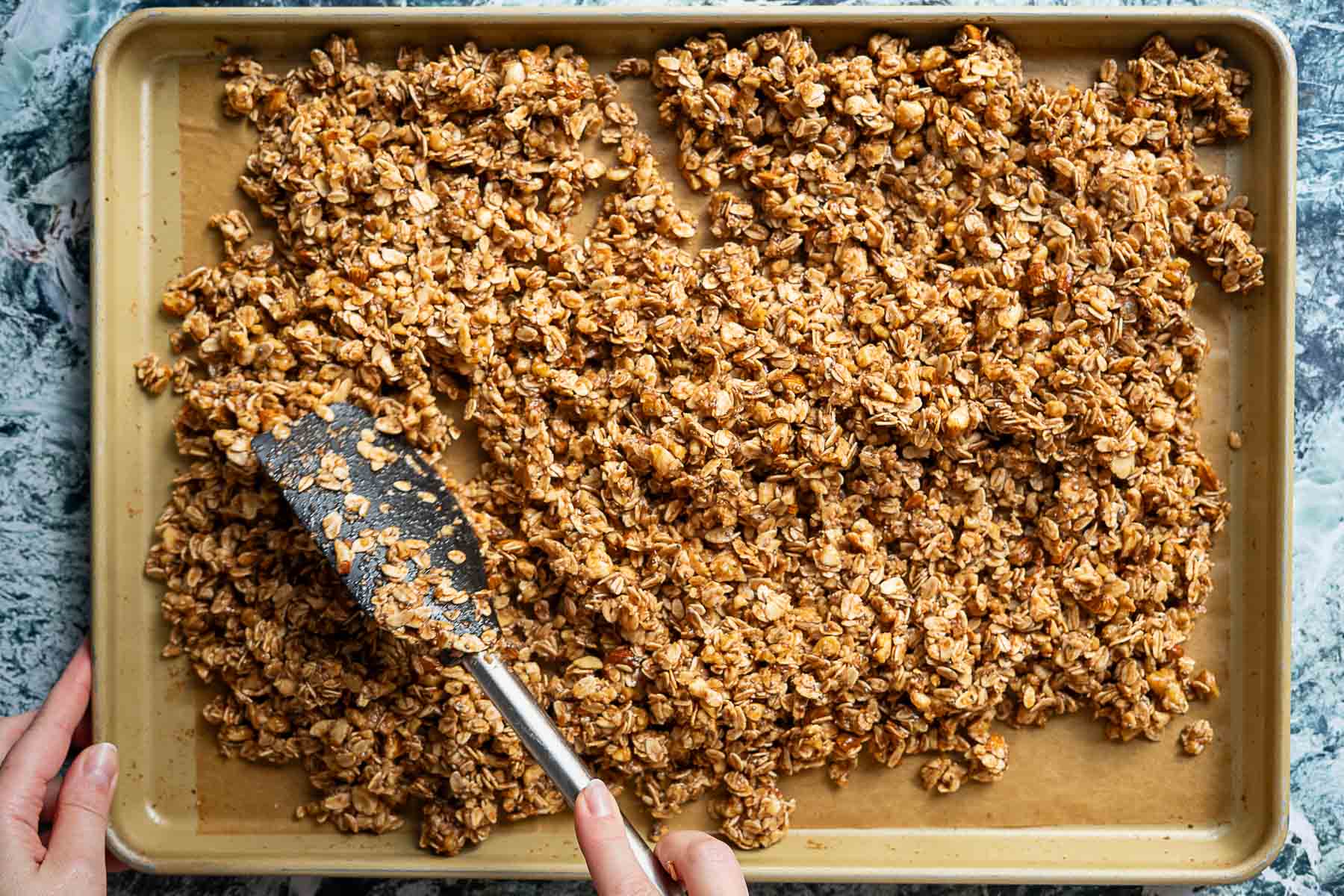 A hand uses a spatula to spread sourdough discard granola mixture evenly on a parchment-lined baking sheet, preparing it for baking.